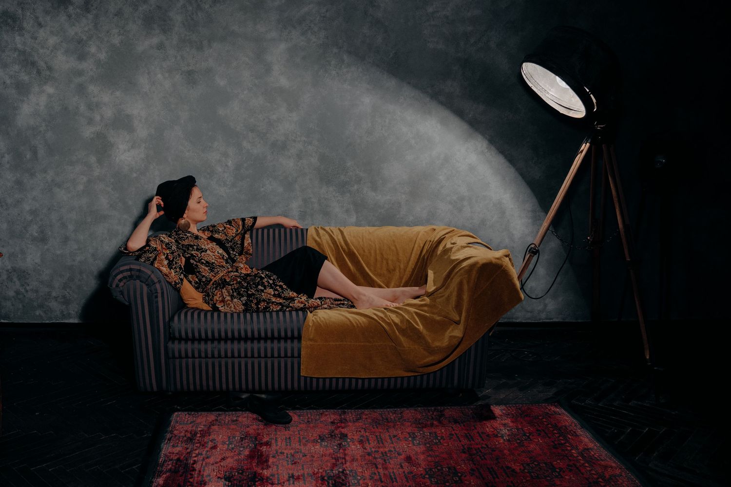 Woman reclining on a mustard-draped sofa under dramatic studio lighting on a dark textured background.