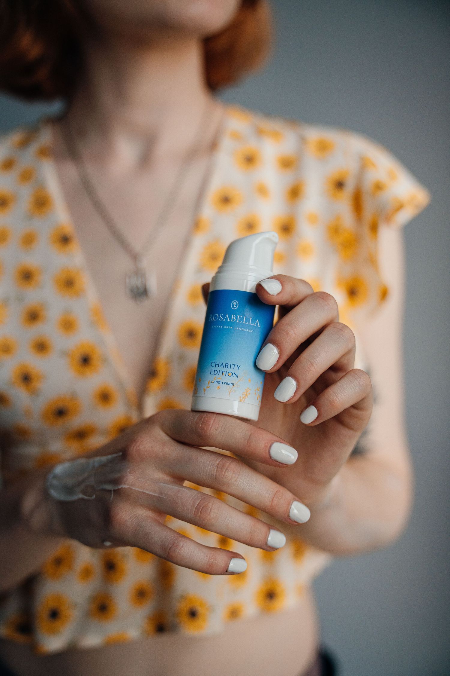 Woman in floral yellow top holding Rosabella Charity Edition facial cream bottle with white manicured nails.
