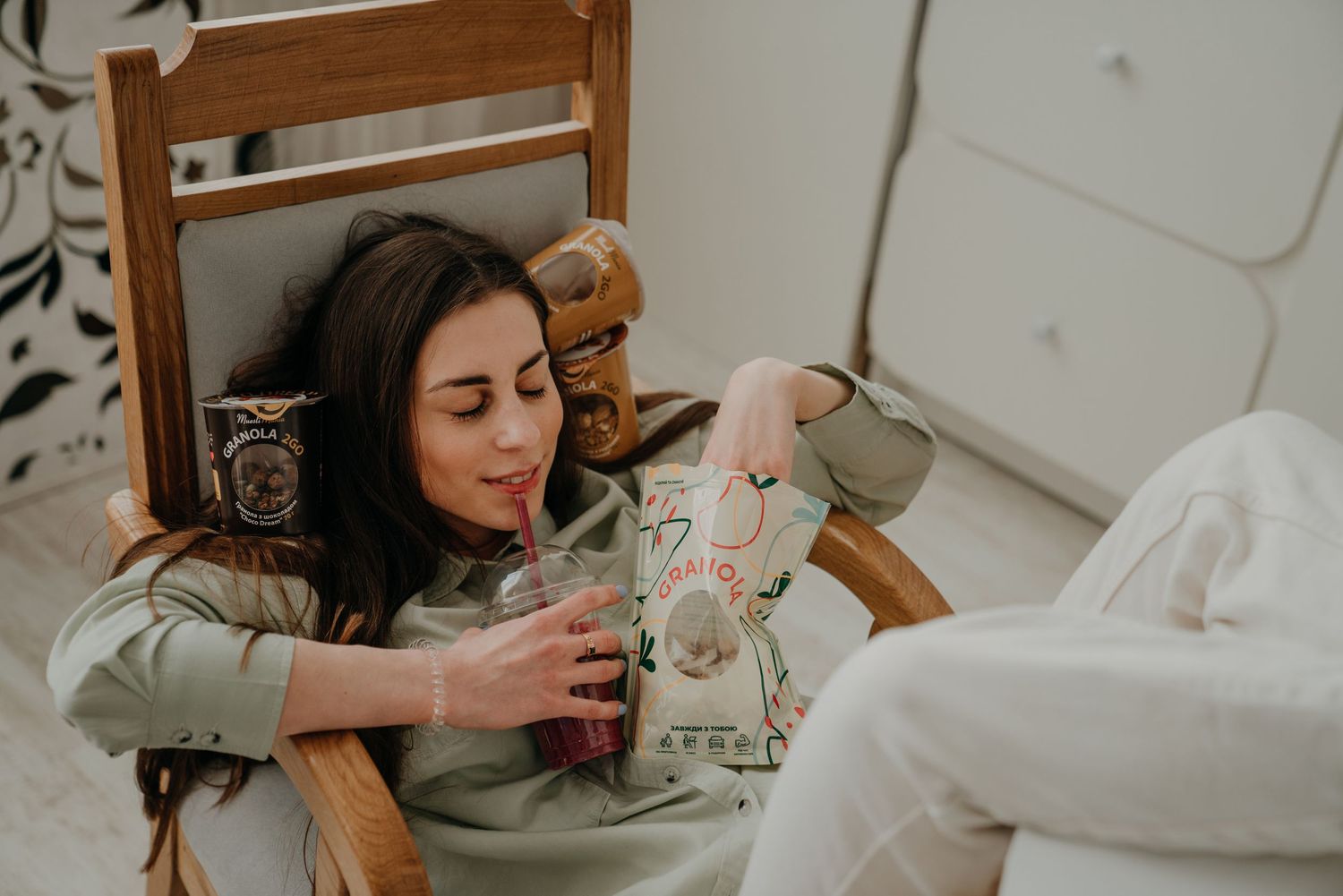 Young woman relaxing in wooden chair, sipping pink drink through straw, wearing sage green hoodie and floral pants.