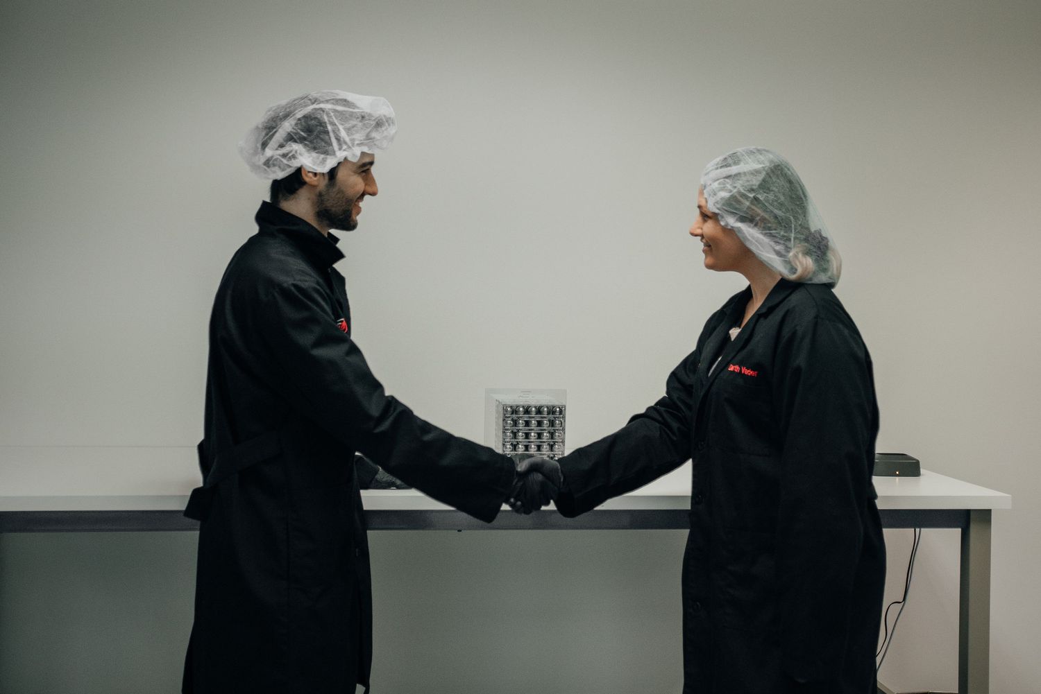 Two food industry workers in black uniforms and hair nets shaking hands in a clean facility.