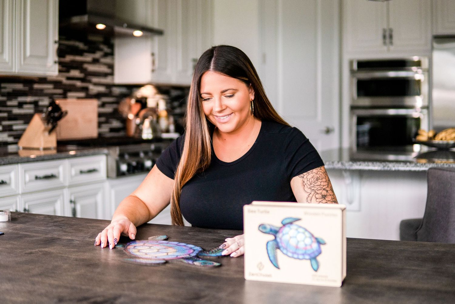 Woman with tattoos smiling while arranging ocean-themed coasters next to a sea turtle art block in a modern kitchen.