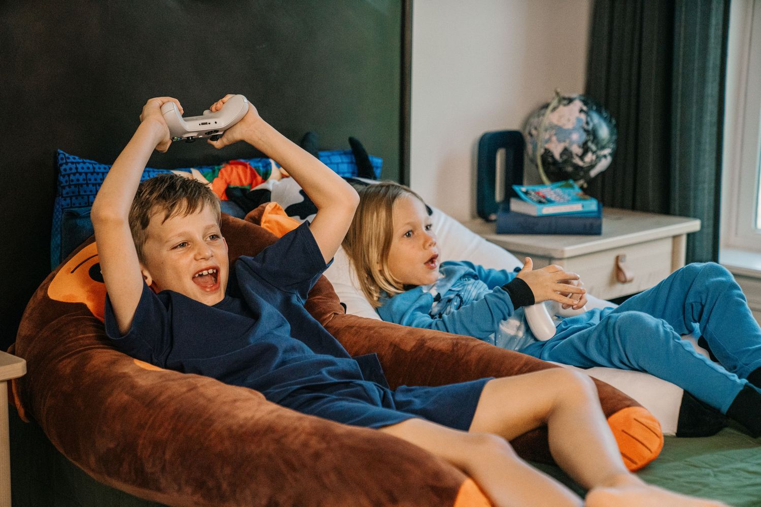 Two excited kids playing video games on brown bean bag chair in bedroom.