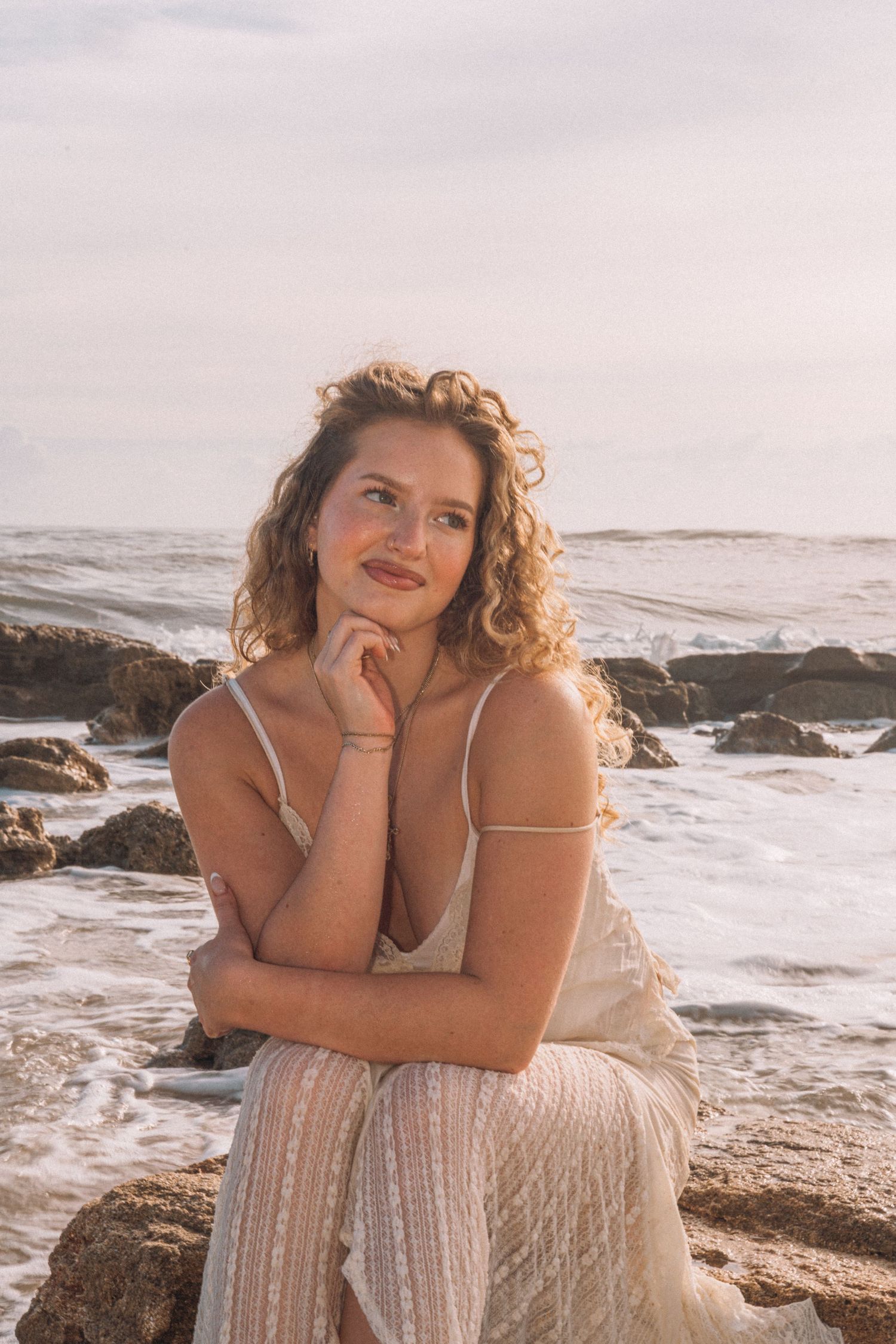 Curly-haired woman in white dress sitting on rocks by the ocean at sunset.
