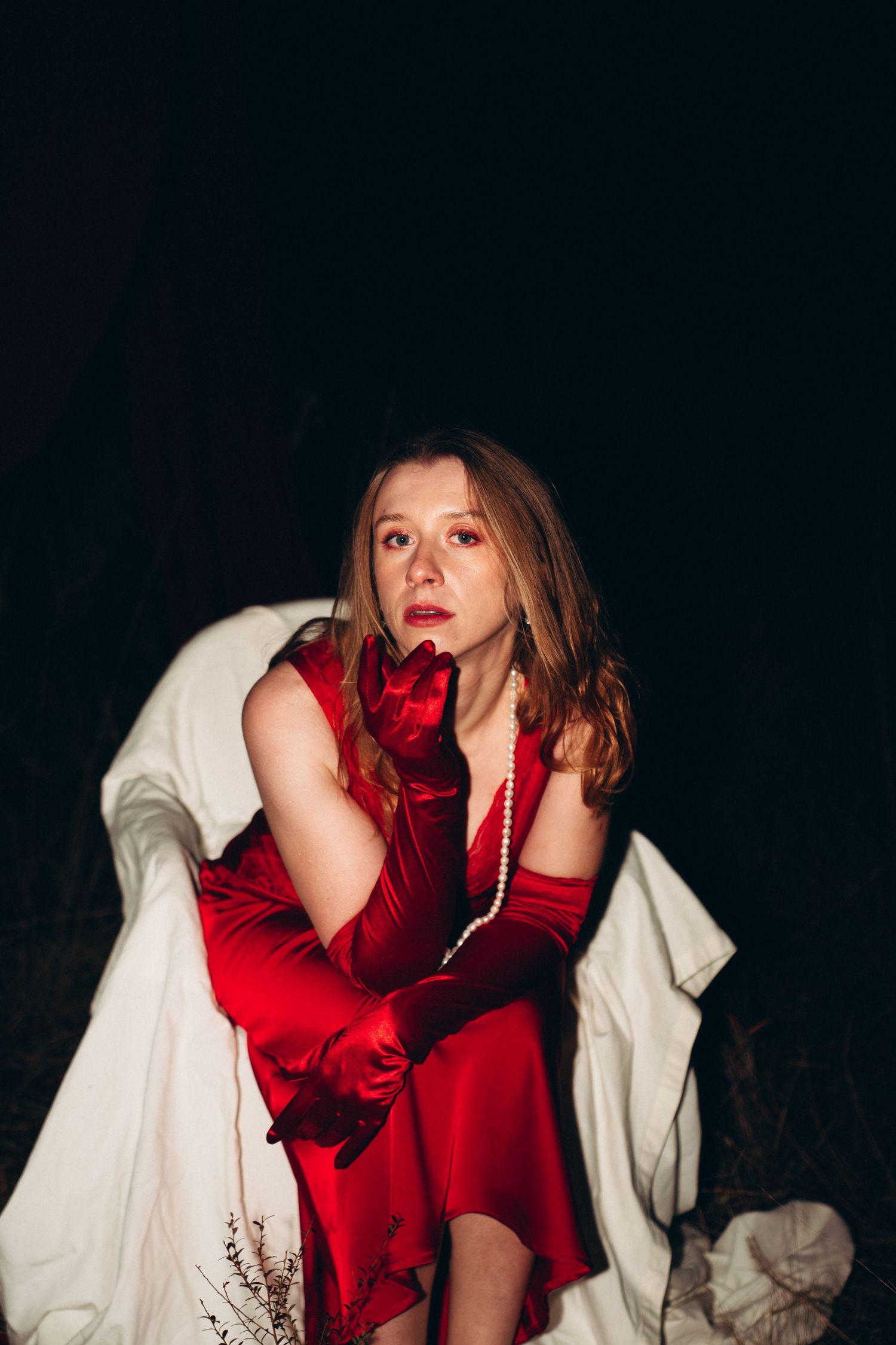 Woman in red satin dress and gloves with pearl necklace poses dramatically against dark background.