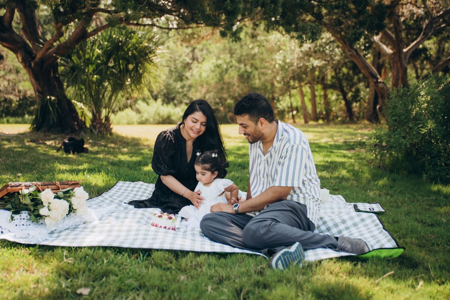 Family enjoying a sunny picnic on a blanket in a lush green park, with a young child between two smiling parents.
