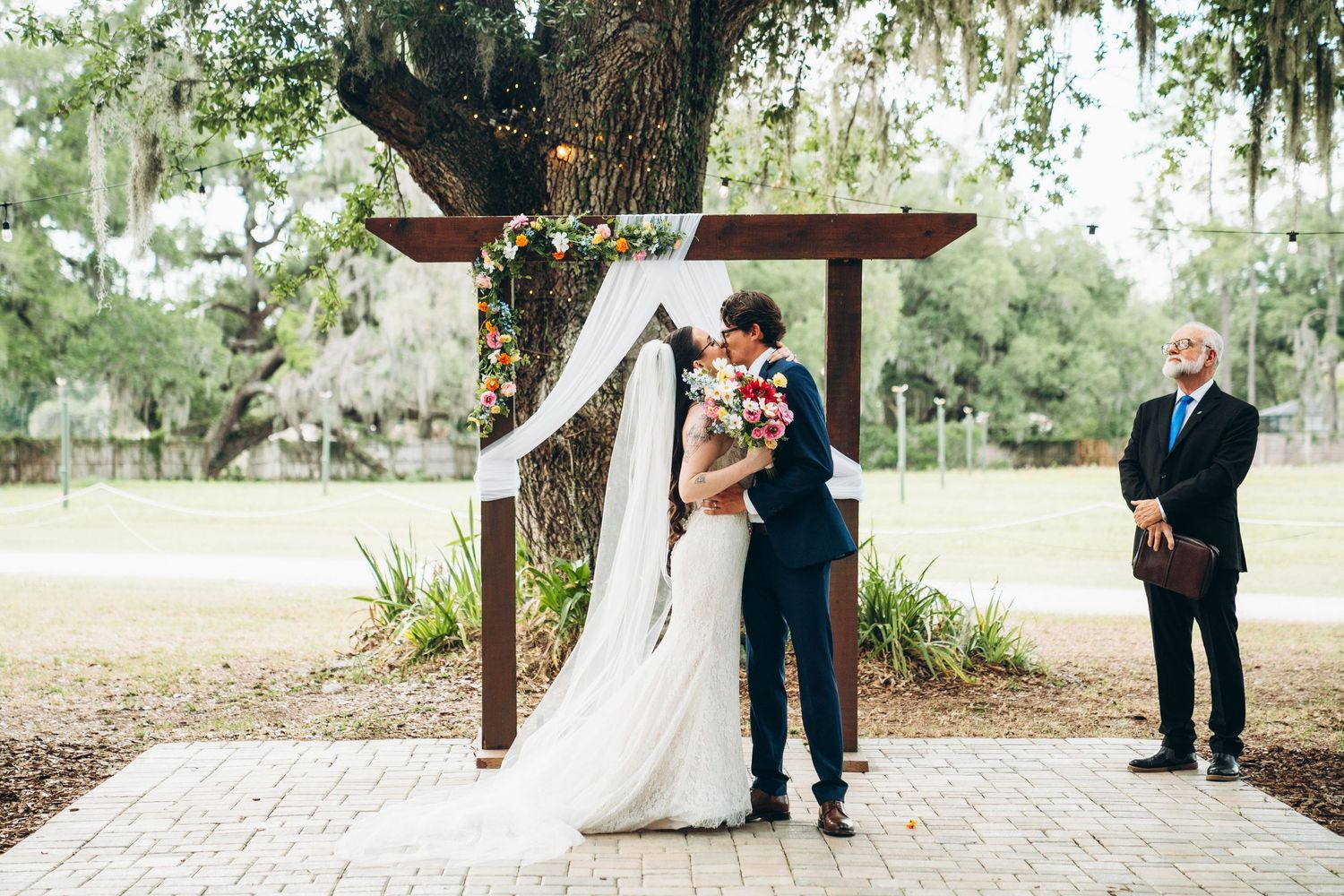 Bride and groom share first kiss under rustic wooden arch adorned with floral decor at outdoor wedding ceremony.