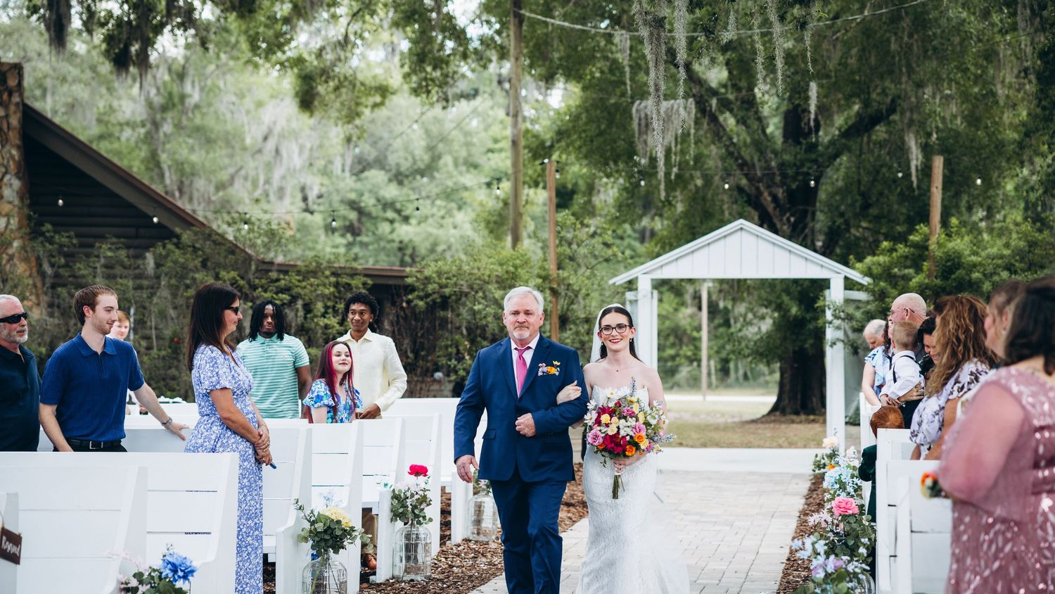 Father walks bride down outdoor aisle at rustic wedding ceremony with white pews and lush green trees.