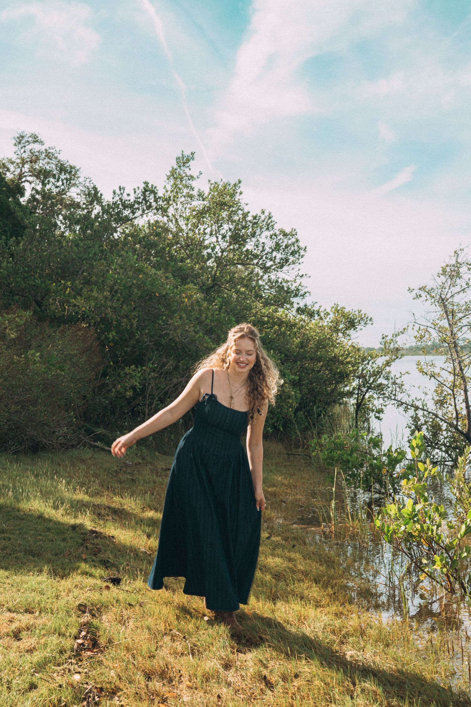A joyful figure in a navy blue dress stands on a grassy hillside against a bright sky.