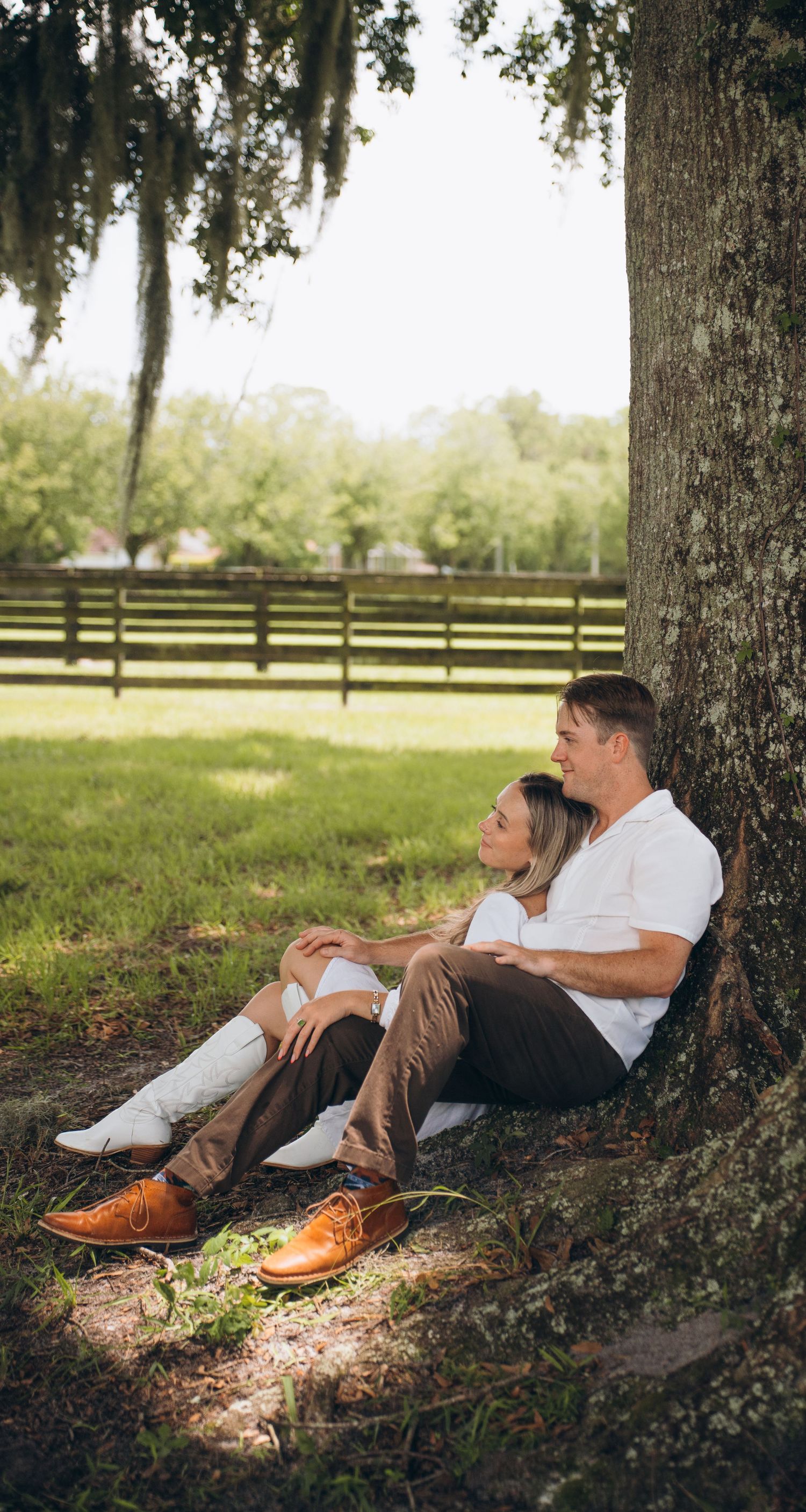 A couple relaxes against a large oak tree in a peaceful pasture setting.