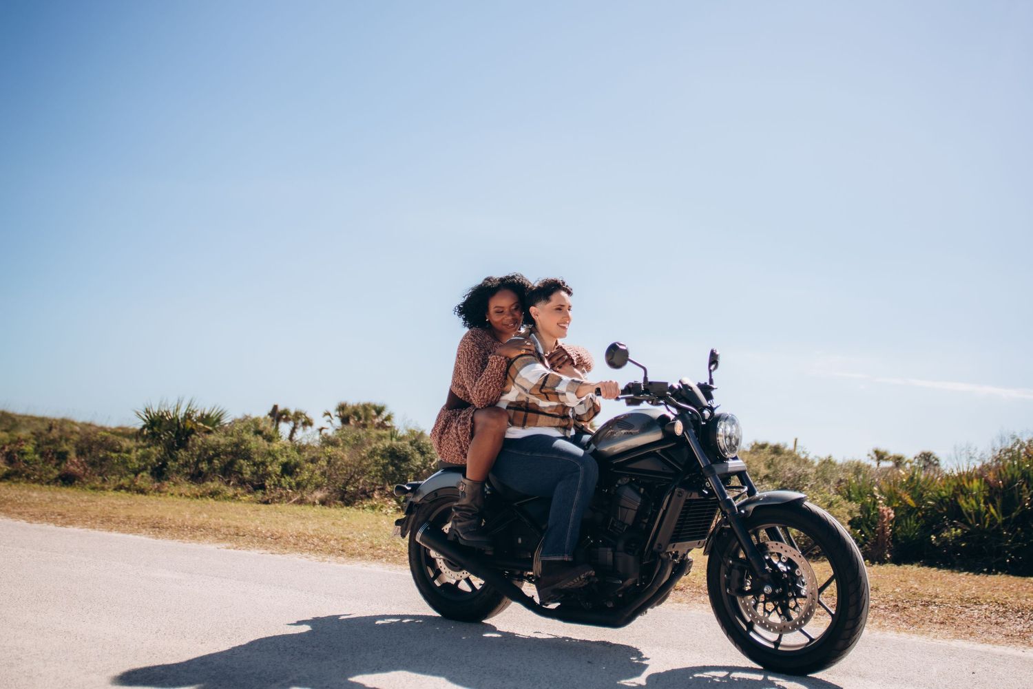 Two people ride together on a black motorcycle against a bright blue sky with palm trees.