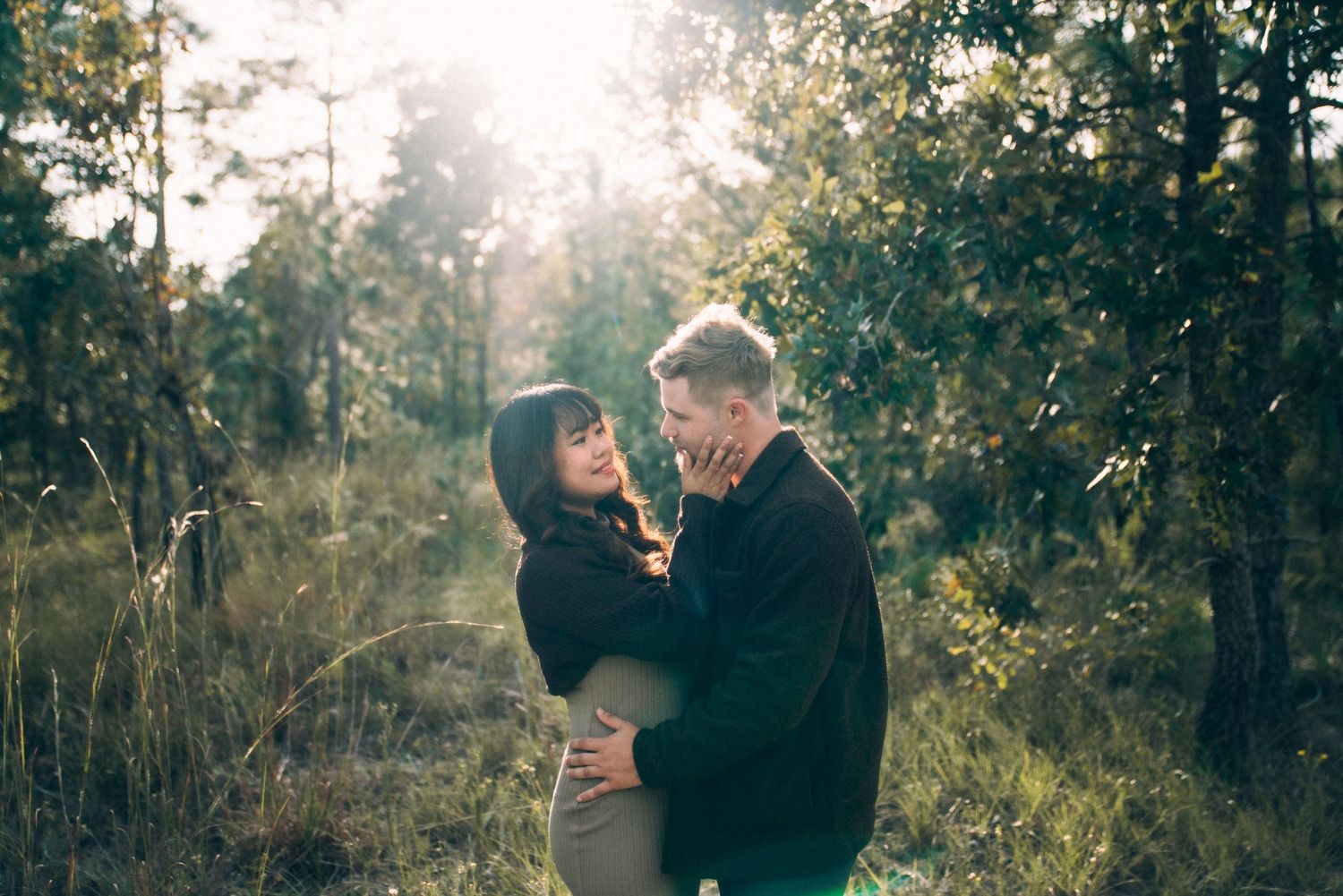 Romantic couple embraces in a misty forest setting during golden hour with sunlight filtering through trees.