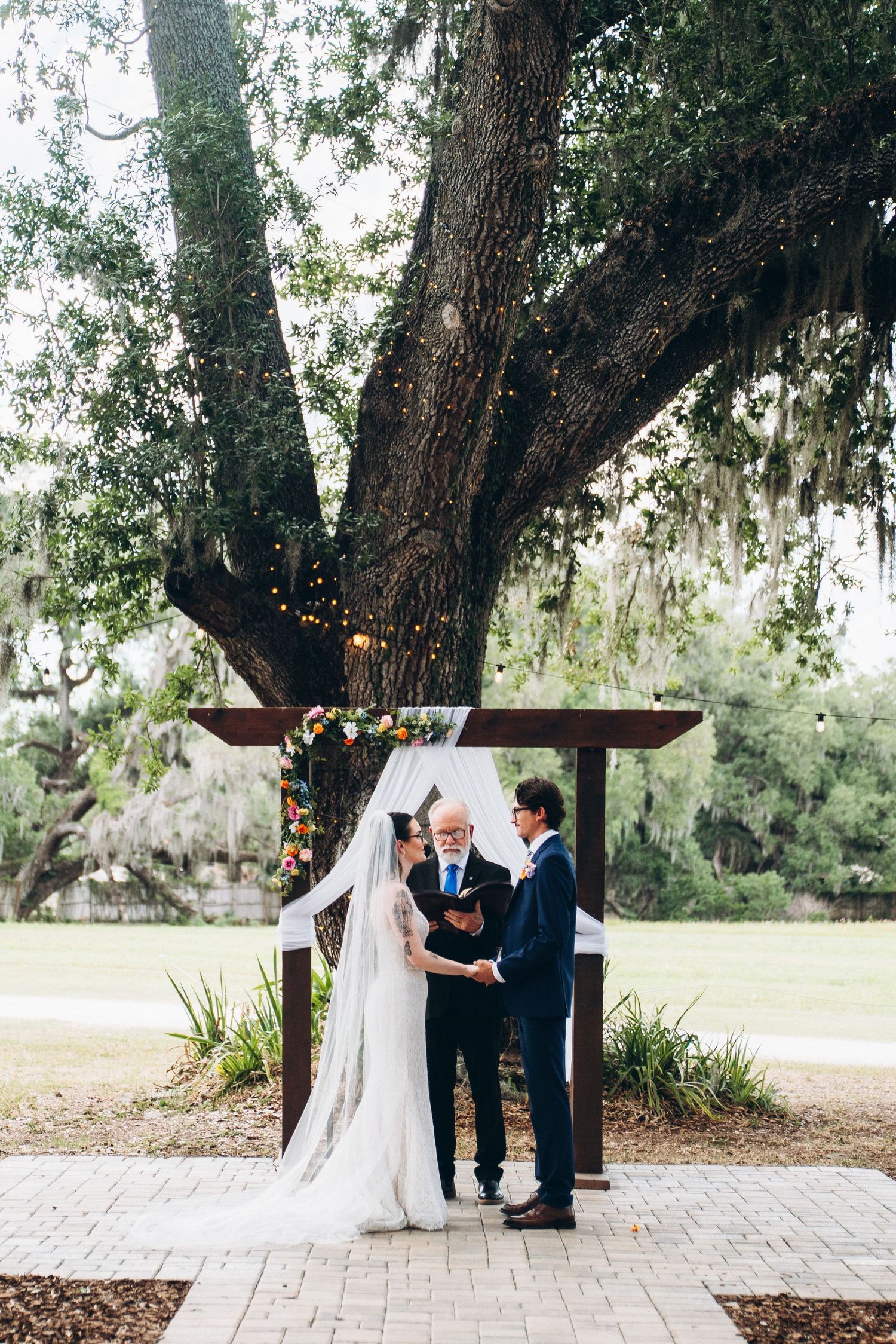 Outdoor wedding ceremony under a large oak tree with string lights, bride in white gown and groom in navy suit with officiant.