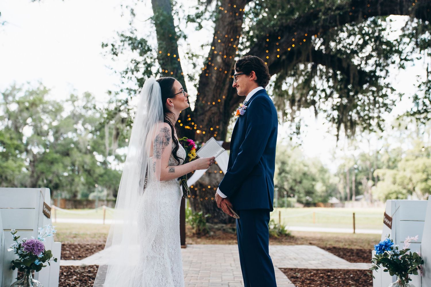 Bride and groom exchange vows outdoors under a large oak tree at an elegant garden wedding ceremony.