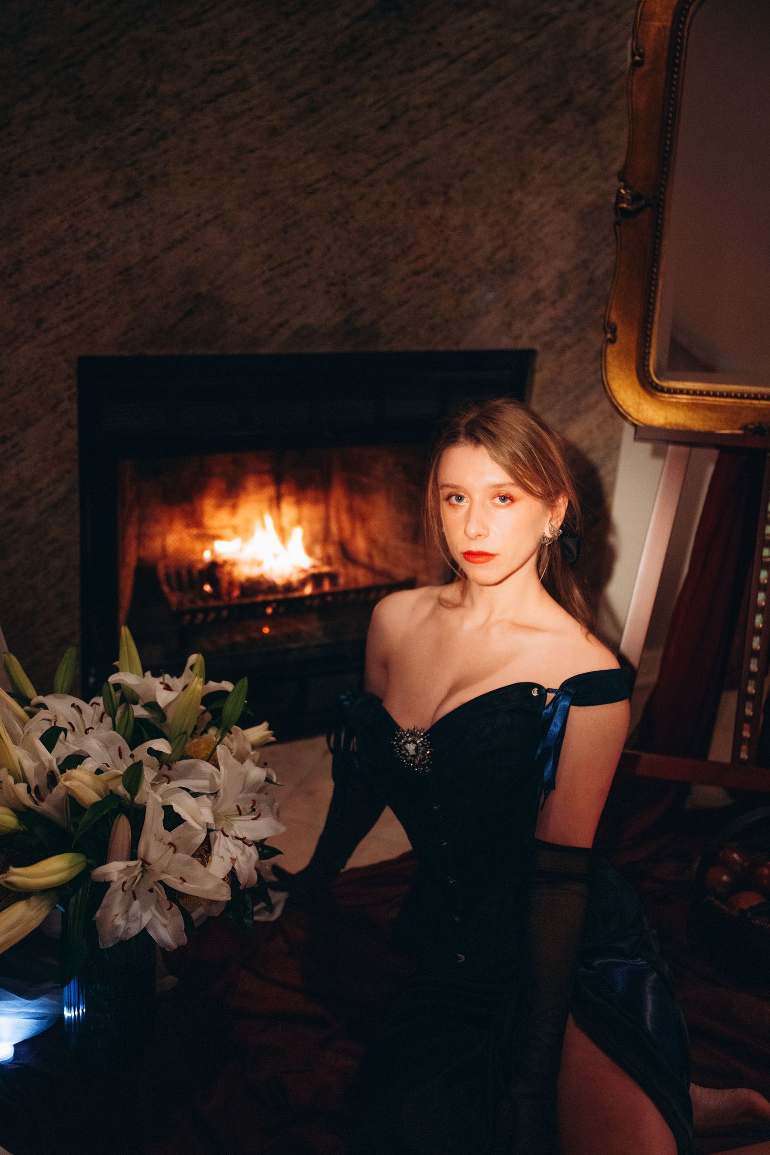 Young woman in elegant black off-shoulder gown sitting by fireplace with white lilies nearby.