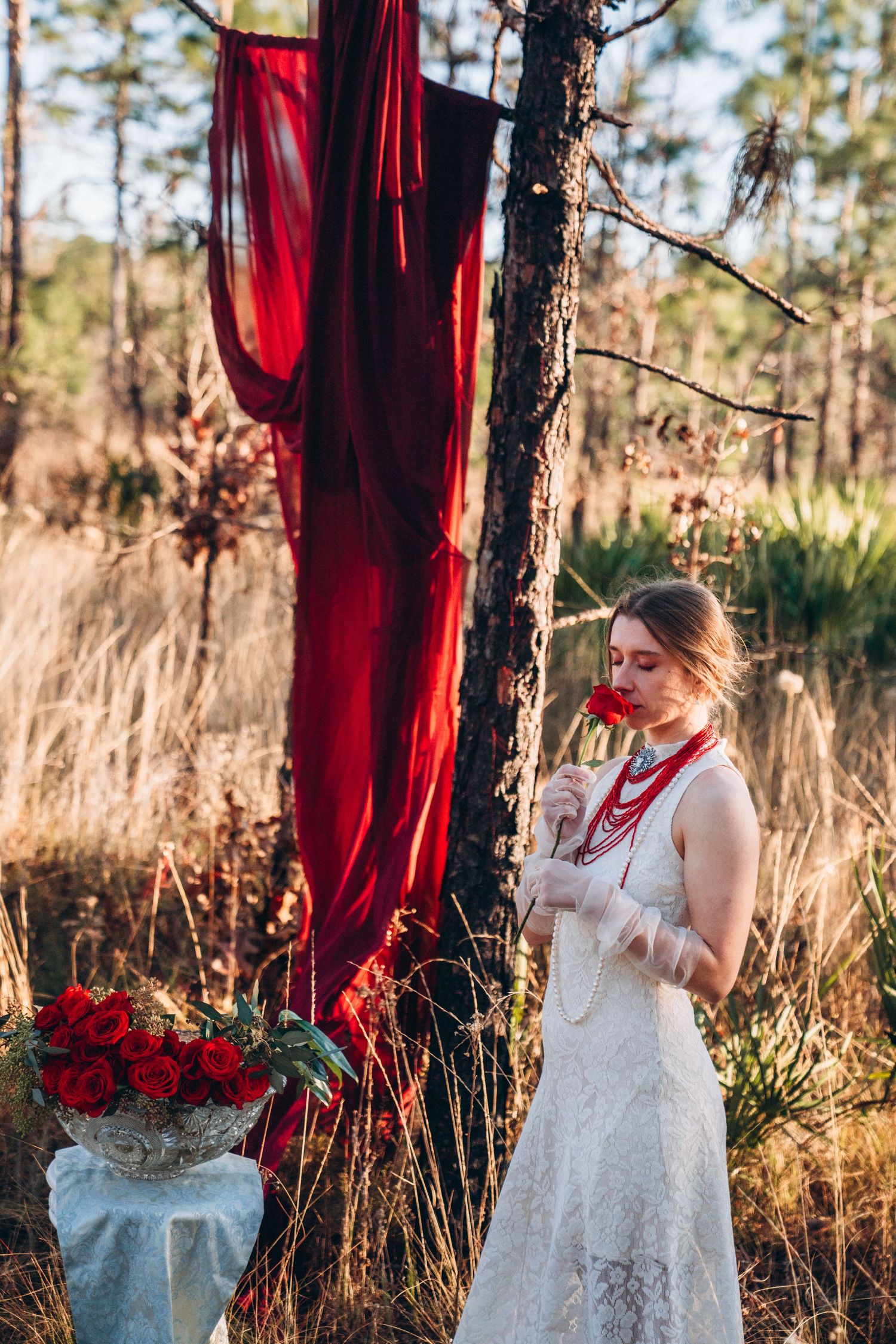 Bride in white lace dress smelling red rose near tree with red fabric draping and bouquet of roses in forest setting.