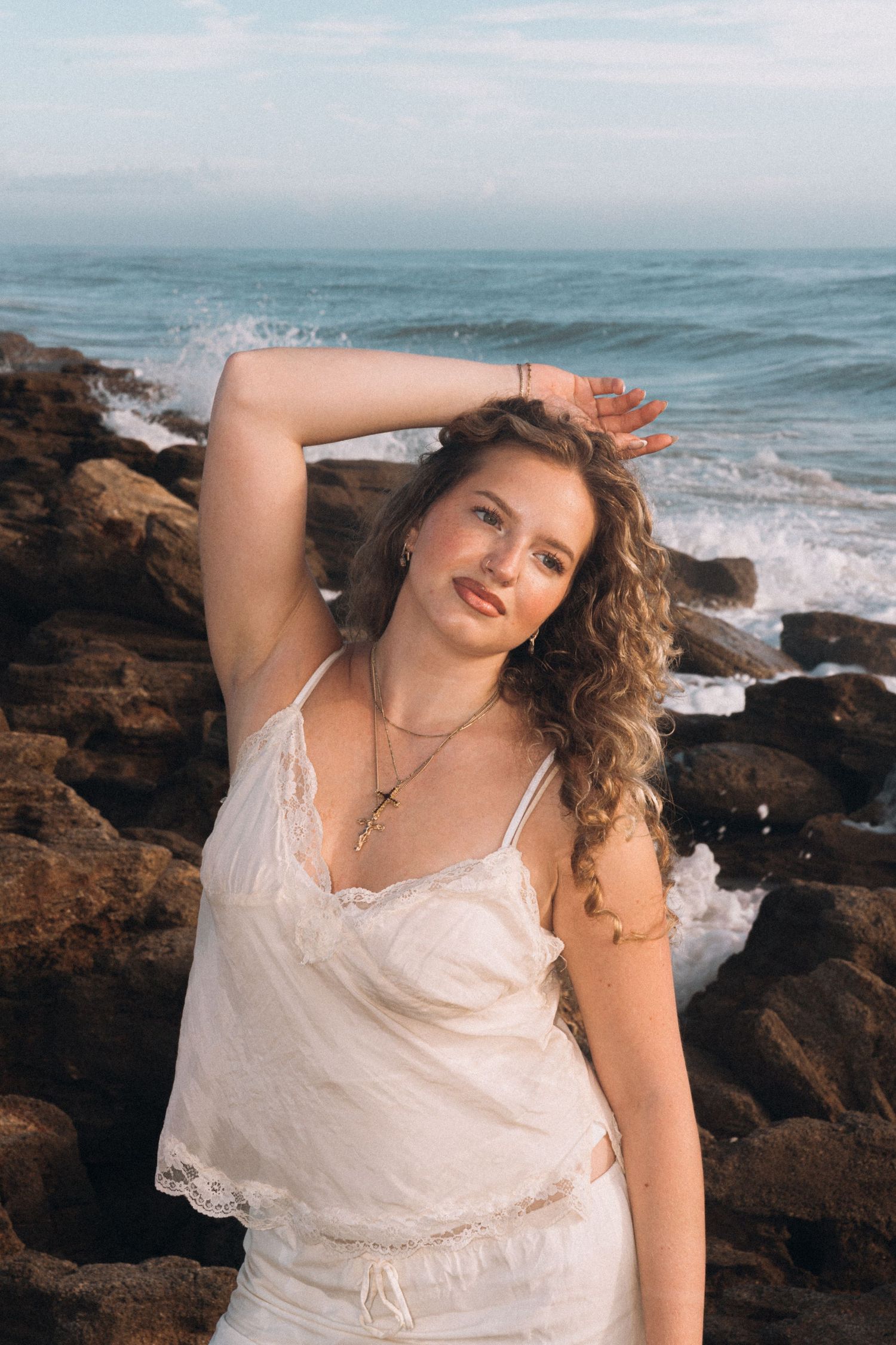 Young woman in white lace dress posing on rocky ocean shore with waves crashing behind her.