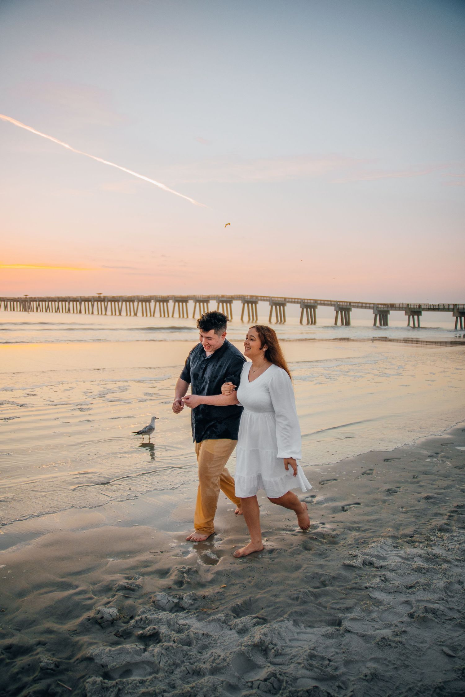 A couple walking along the beach with a long pier visible in the sunset background.