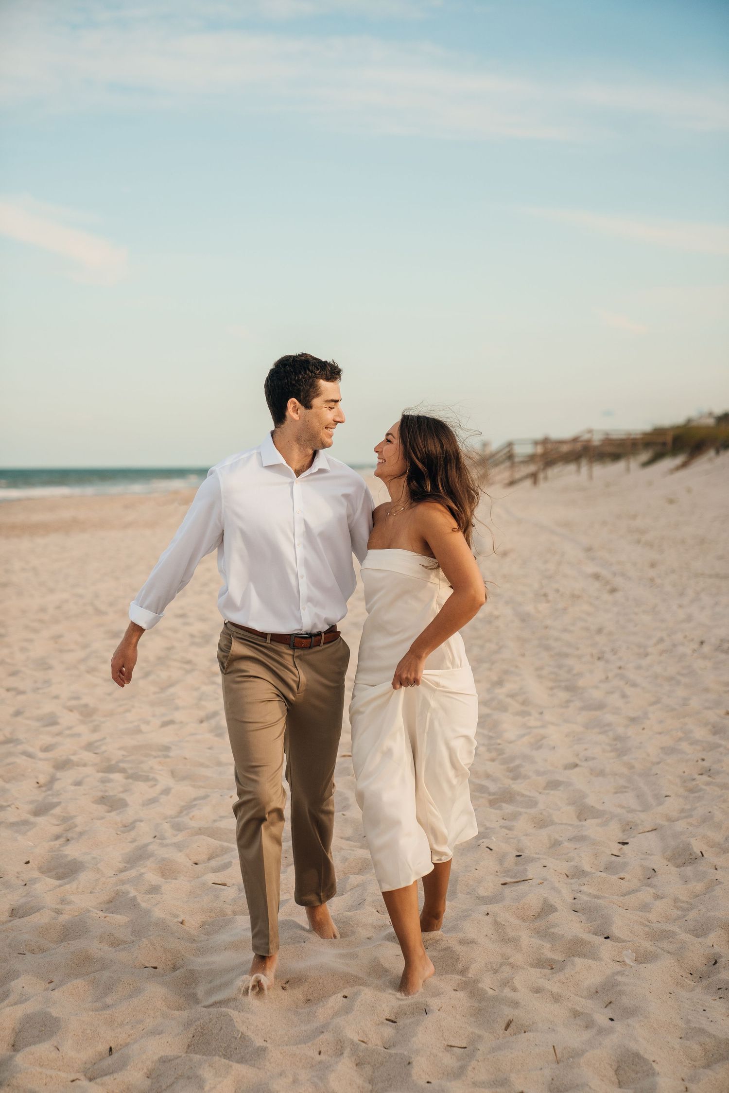 A couple walks barefoot together along a sandy beach shoreline.