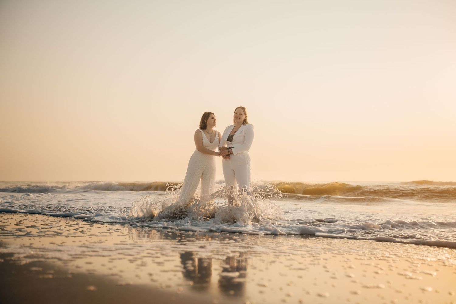 Two people in white outfits stand together on a beach during golden hour.