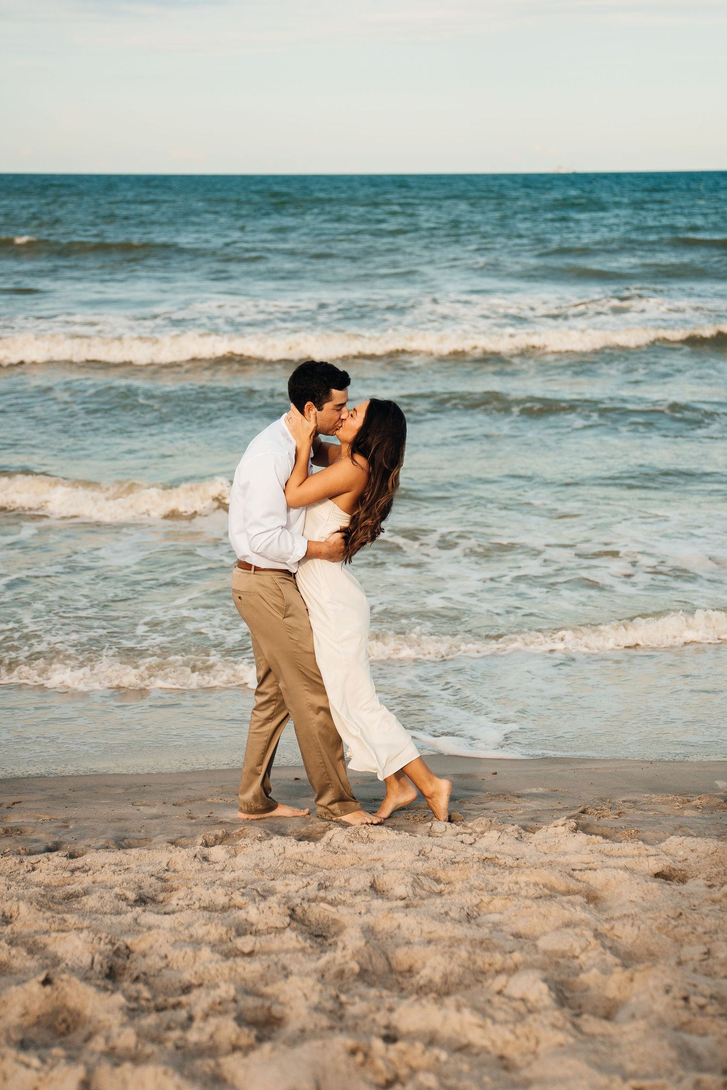 A romantic couple shares an intimate dance together on a sandy beach with rolling ocean waves in the background.