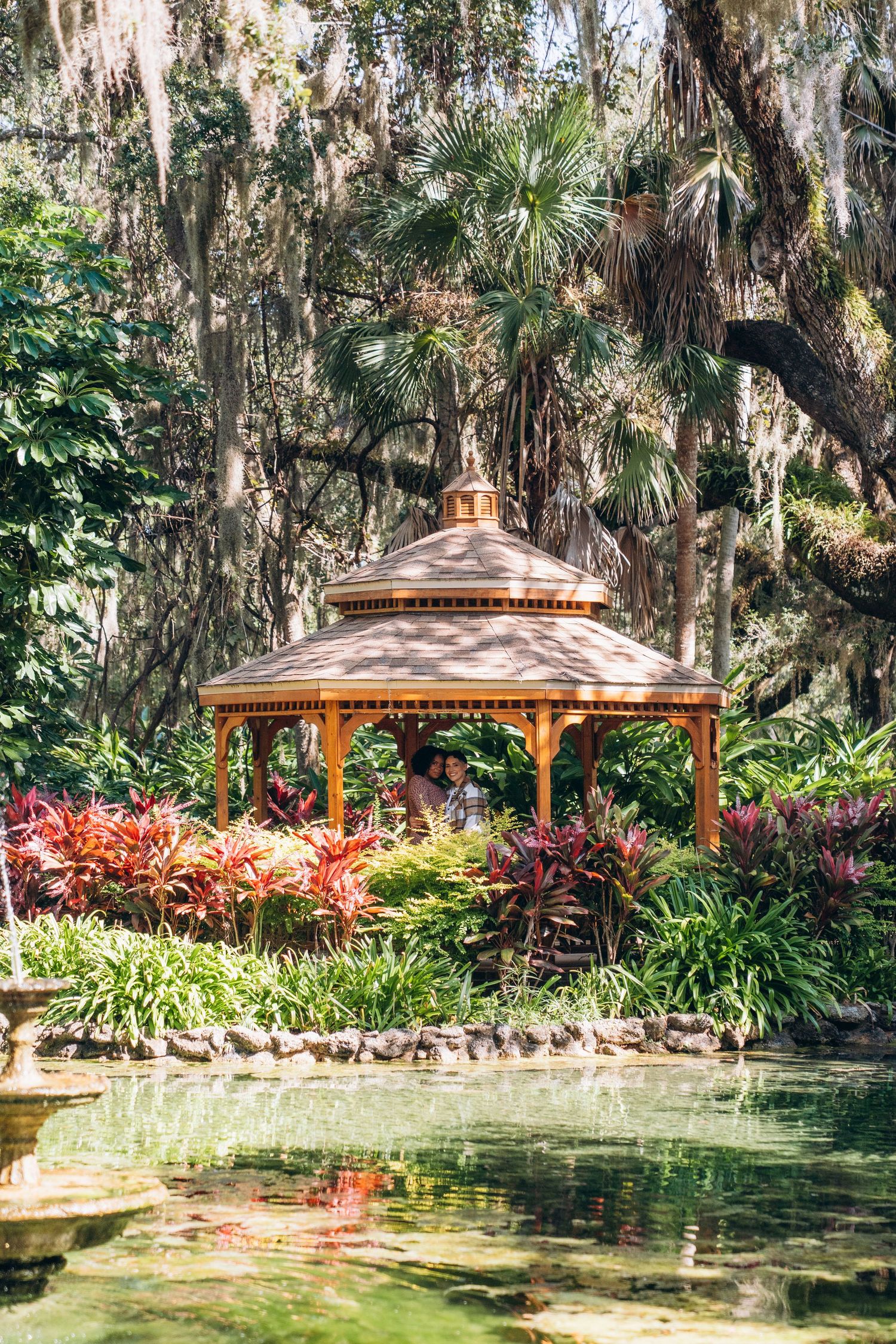 A wooden gazebo sits beside a tranquil pond surrounded by colorful tropical plants and hanging moss.