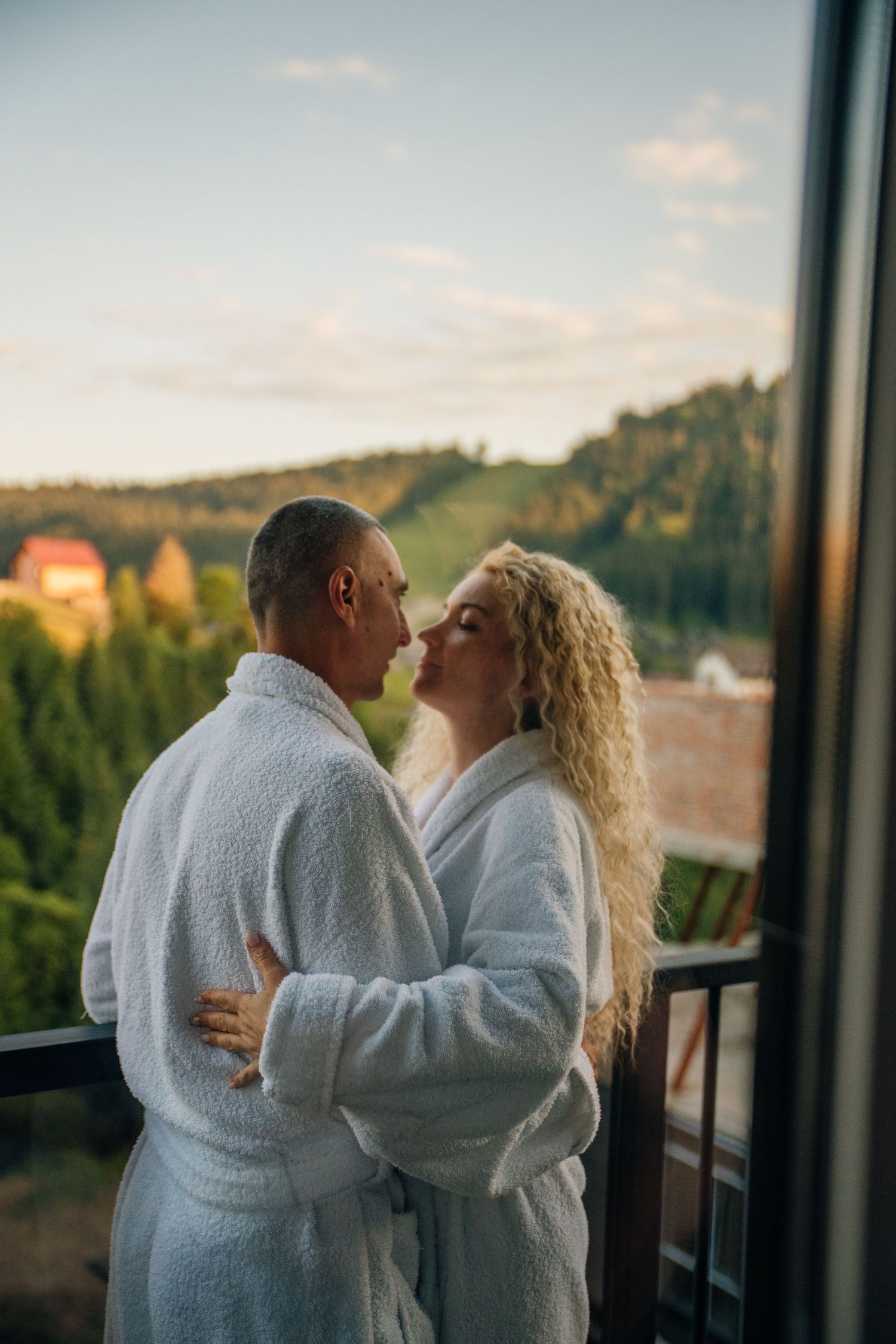 Couple in white bathrobes embracing on a balcony with scenic forest view at sunset.