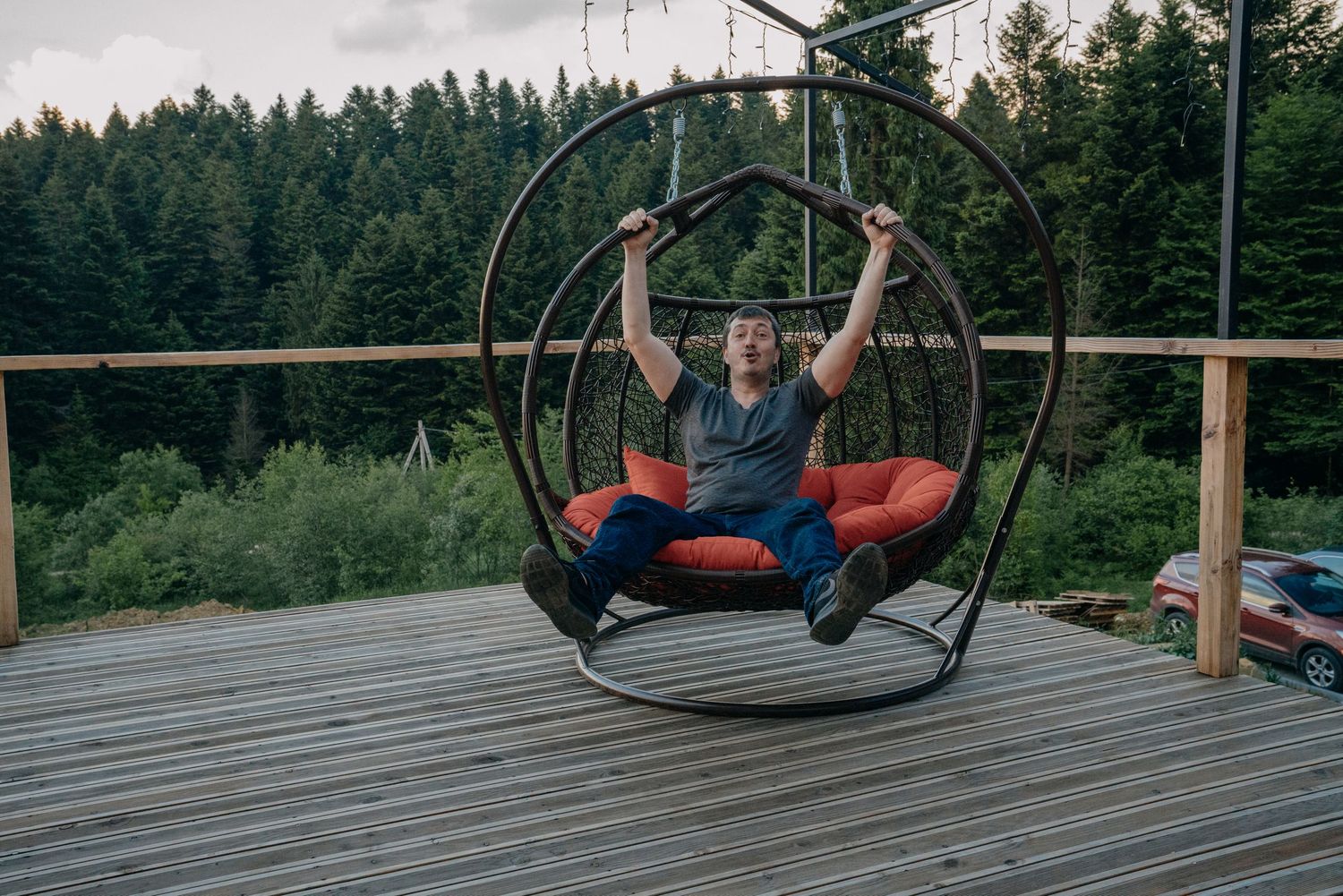 Happy man sitting in hanging bubble chair on wooden deck surrounded by forest.