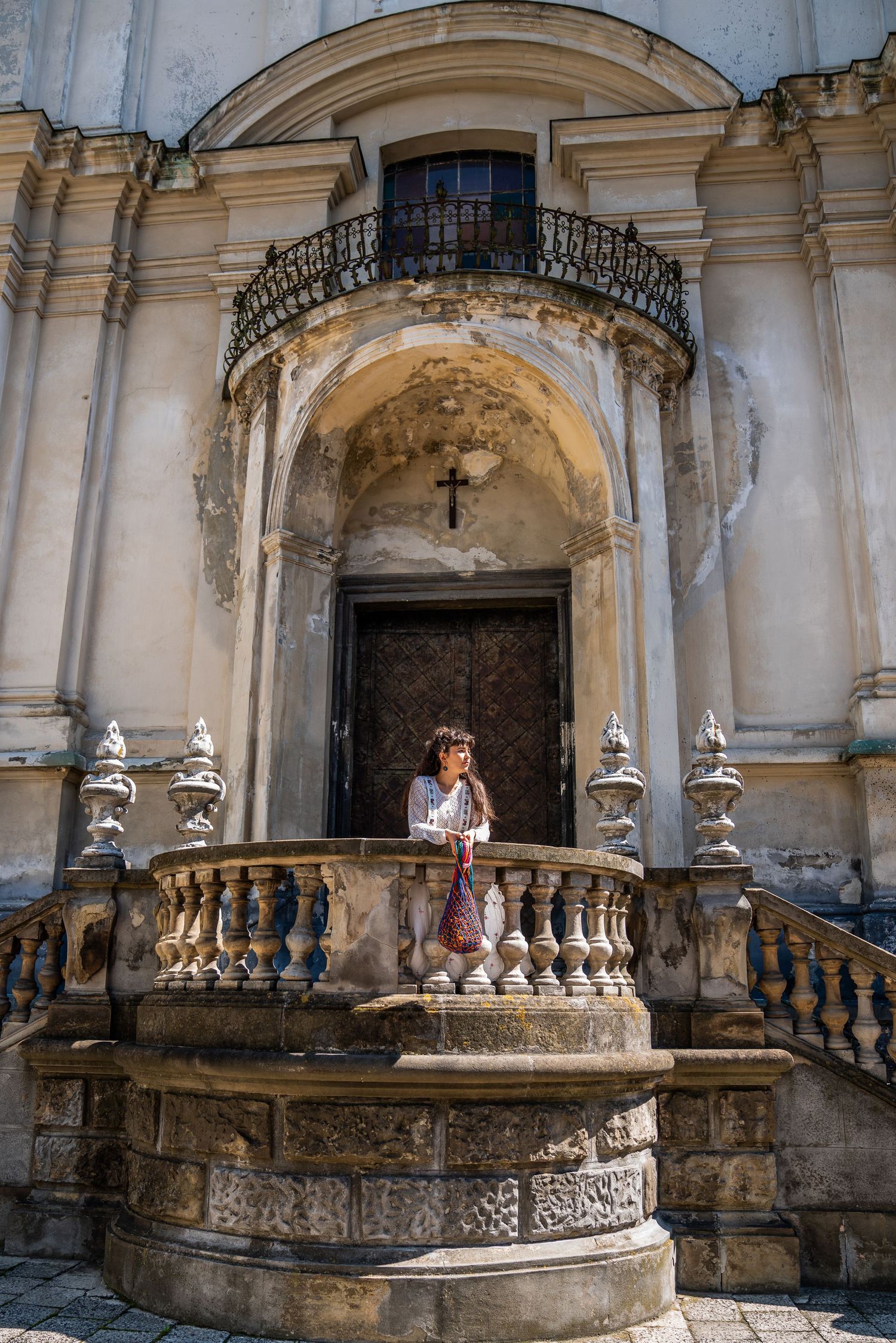Woman standing on ornate stone staircase of historic baroque church with cross above doorway.