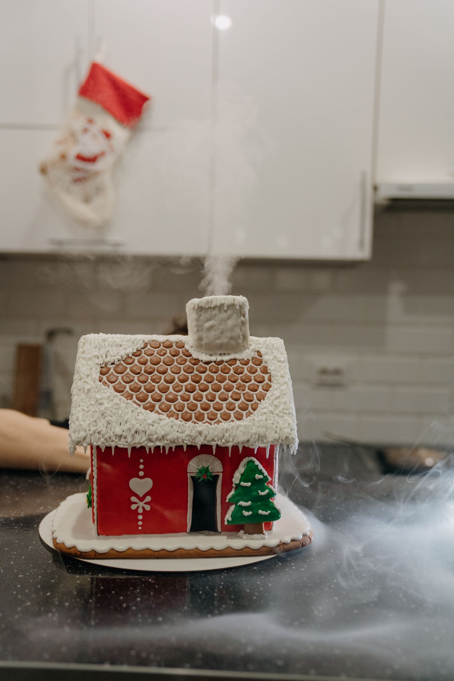 Festive gingerbread house with white icing snow, red walls, and Christmas tree decoration, emitting smoke from chimney in kitchen.