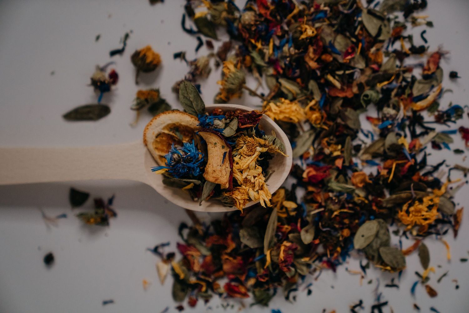 Wooden spoon with colorful dried herbal tea blend of flowers and leaves scattered on white background.