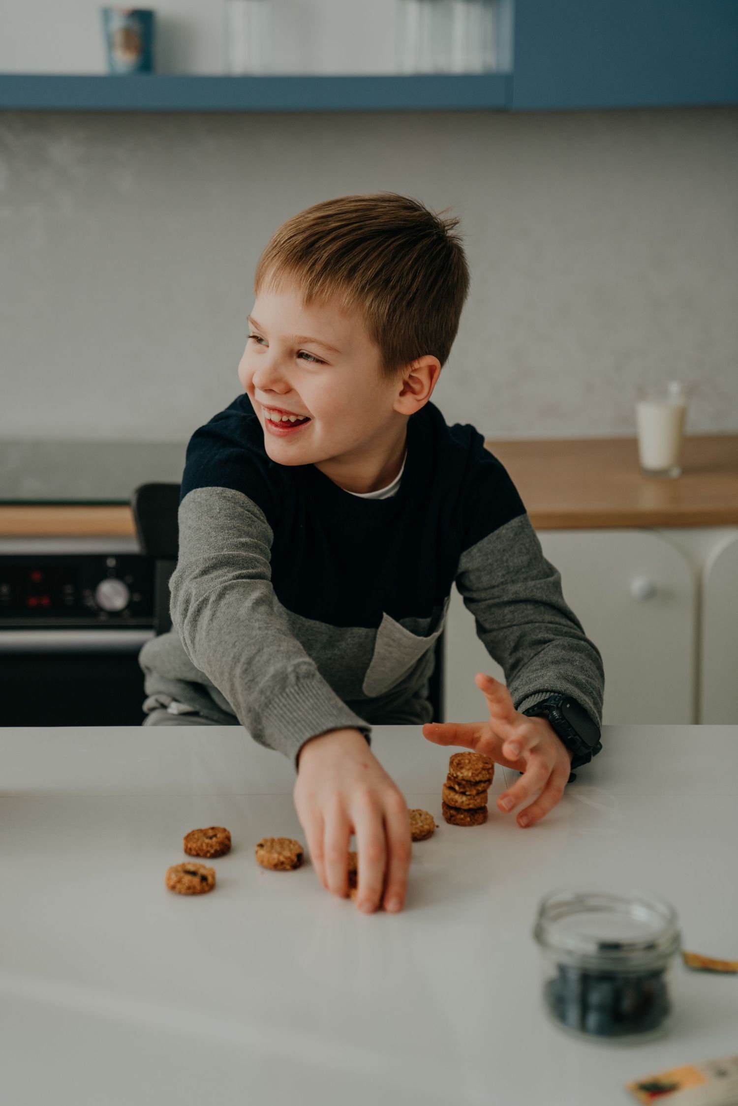 A happy young boy stacking cookies on a white kitchen counter with a glass of milk nearby.