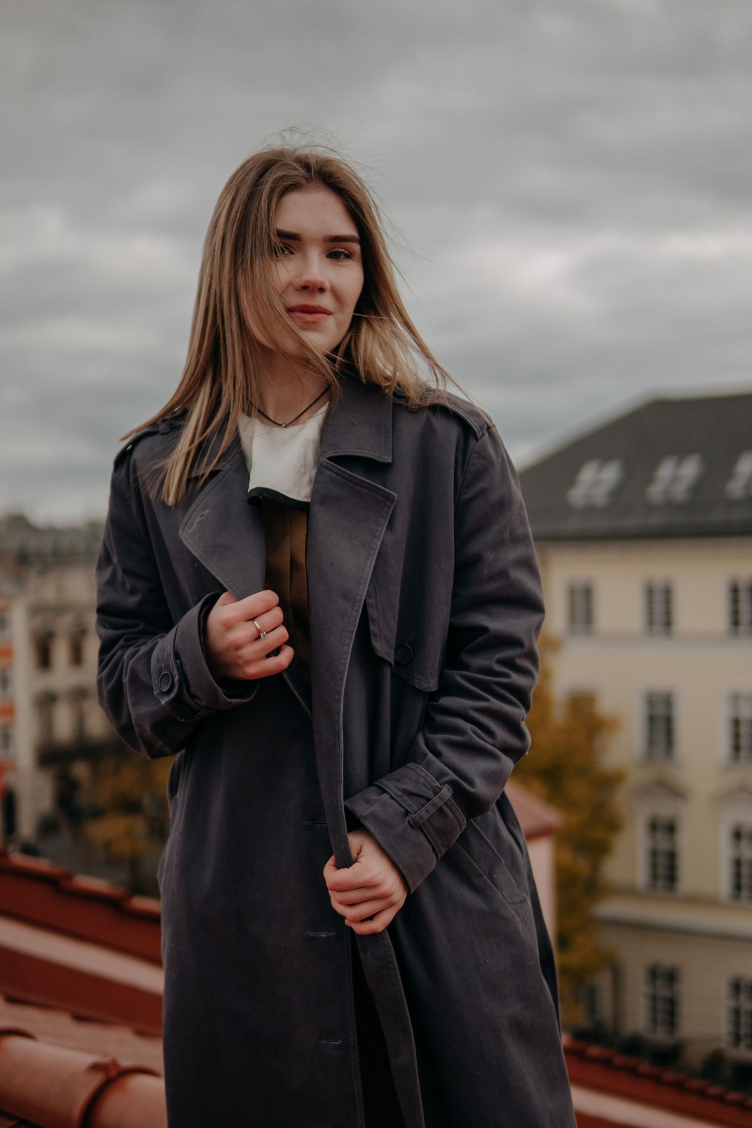 Young woman in gray trench coat posing on rooftop with cloudy sky and European buildings in background.