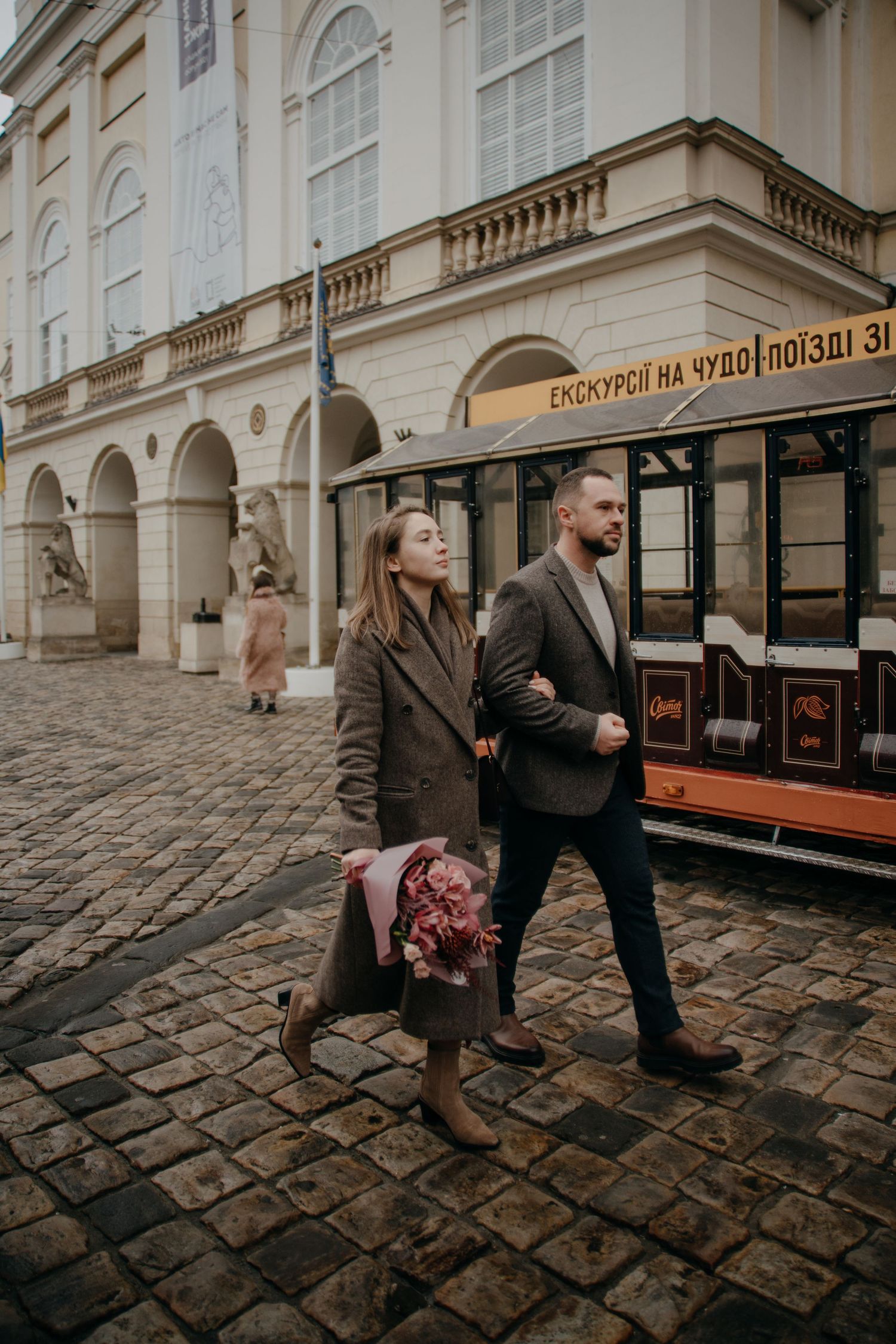 Couple walking on cobblestone street past vintage tram and classical building, woman holding pink bouquet.