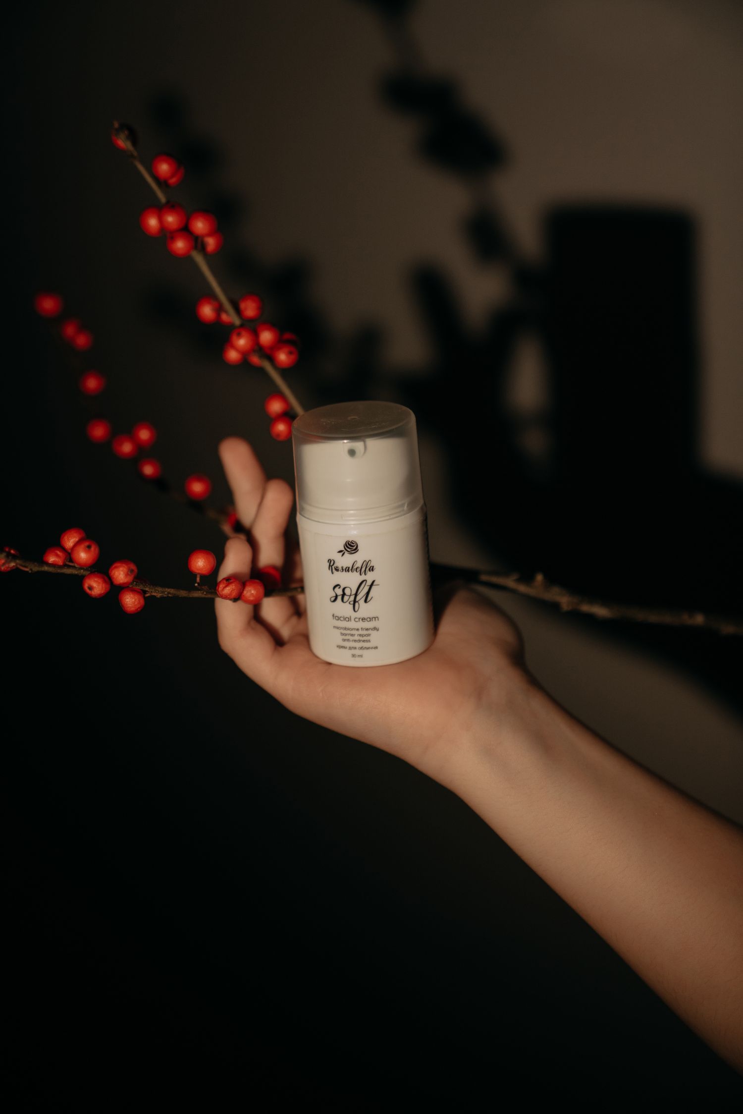 Hand holding a white Beautifin Soft skincare cream bottle near red berry branches in dark setting.