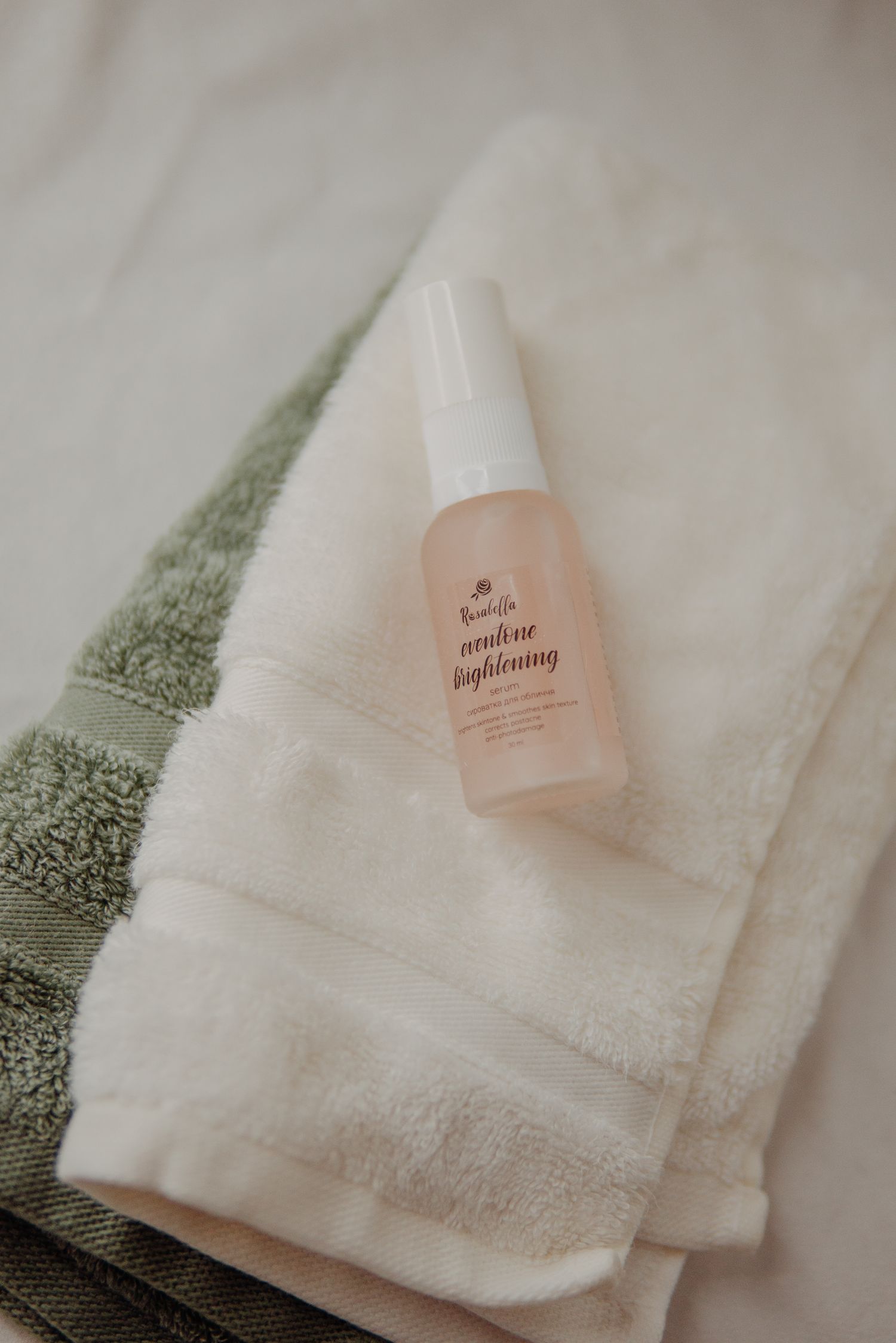 A small pink brightening facial mist spray bottle resting on folded white and green towels.