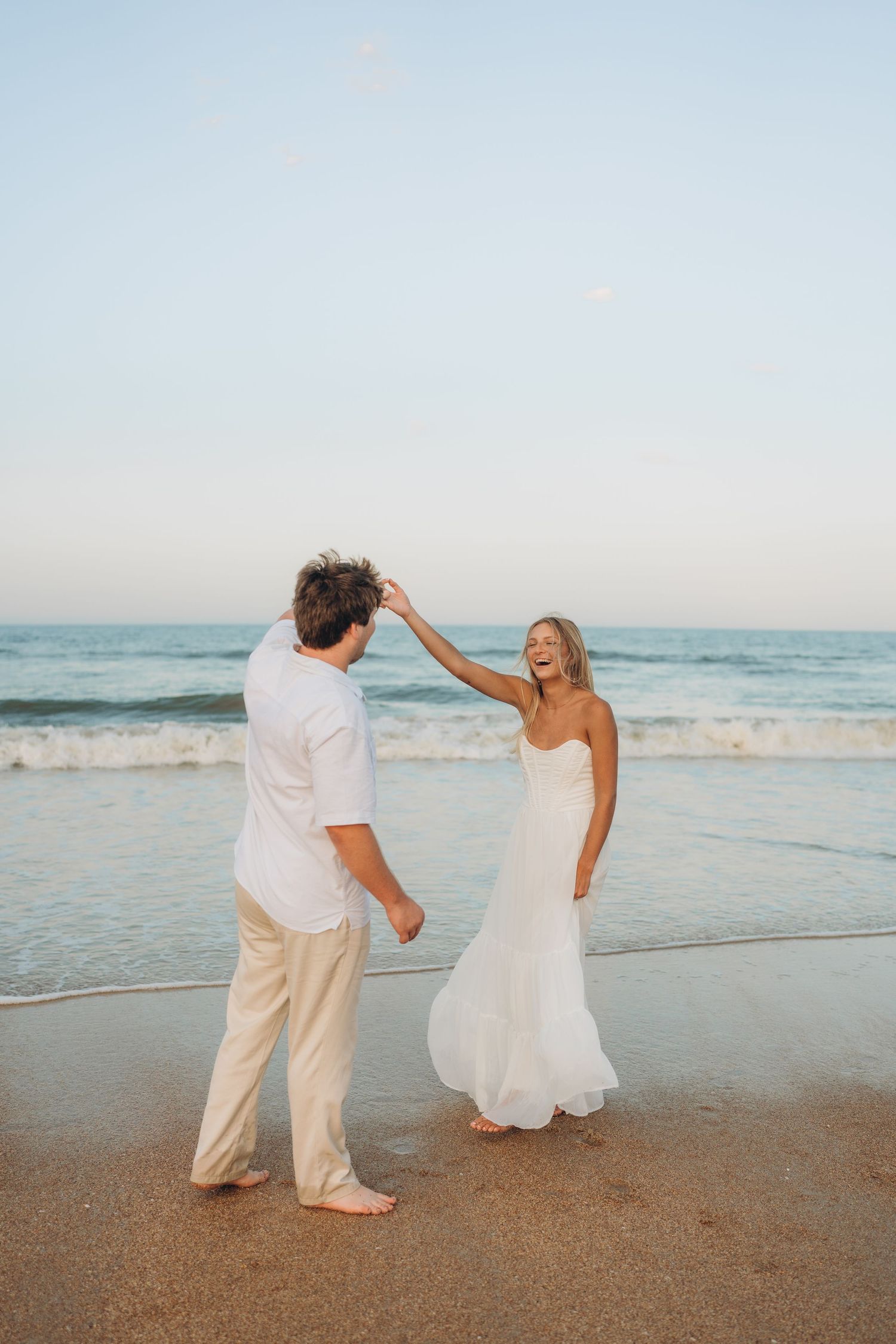 A couple in white outfits dances playfully on the beach with waves crashing behind them.