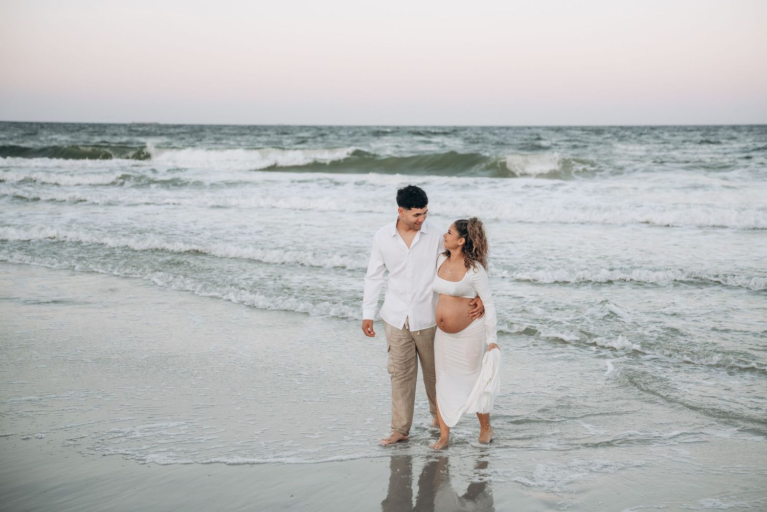 A couple in white attire walks together along the shoreline as waves crash on a cloudy beach.