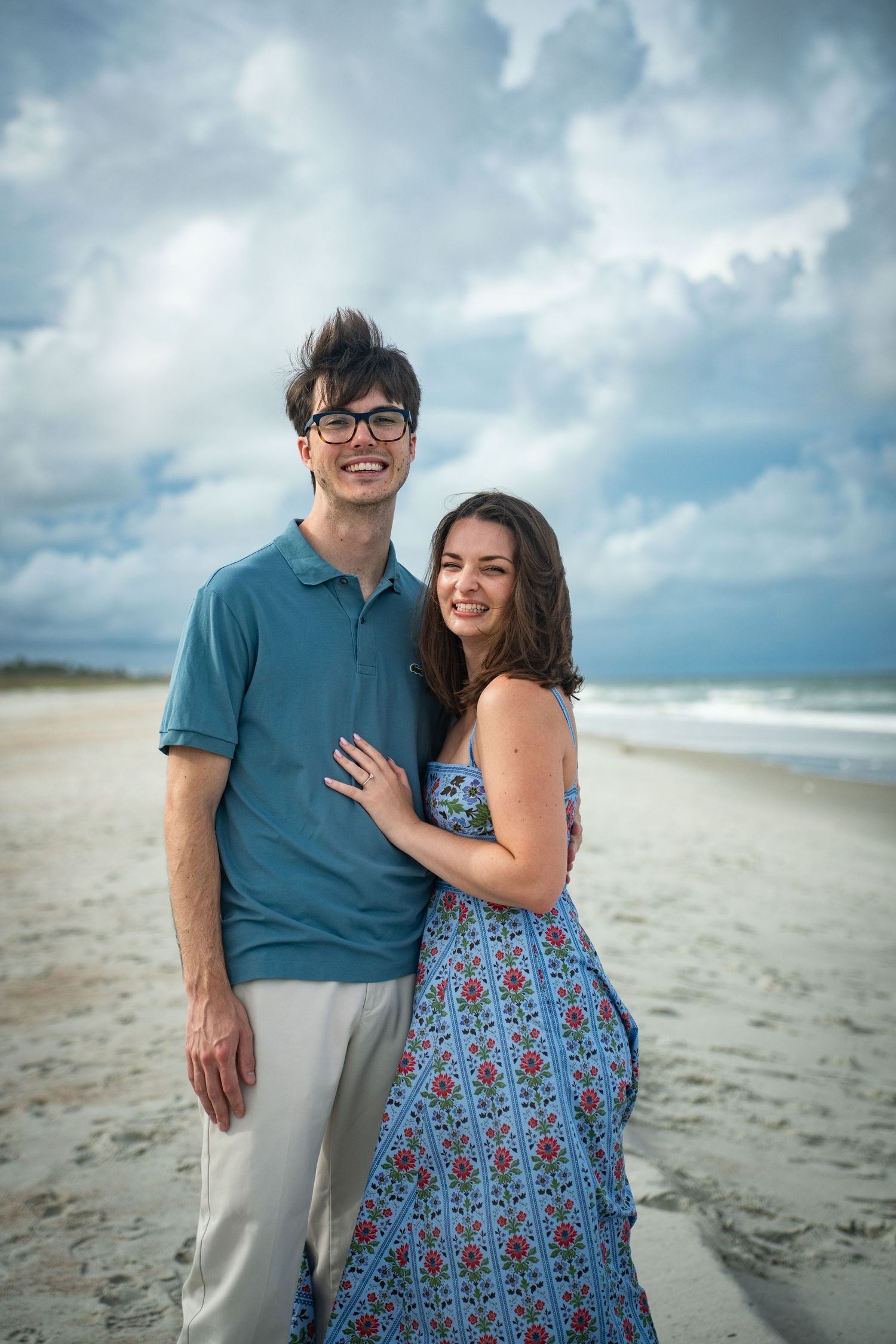 A couple shares a romantic moment together on a cloudy beach wearing blue and floral print outfits.