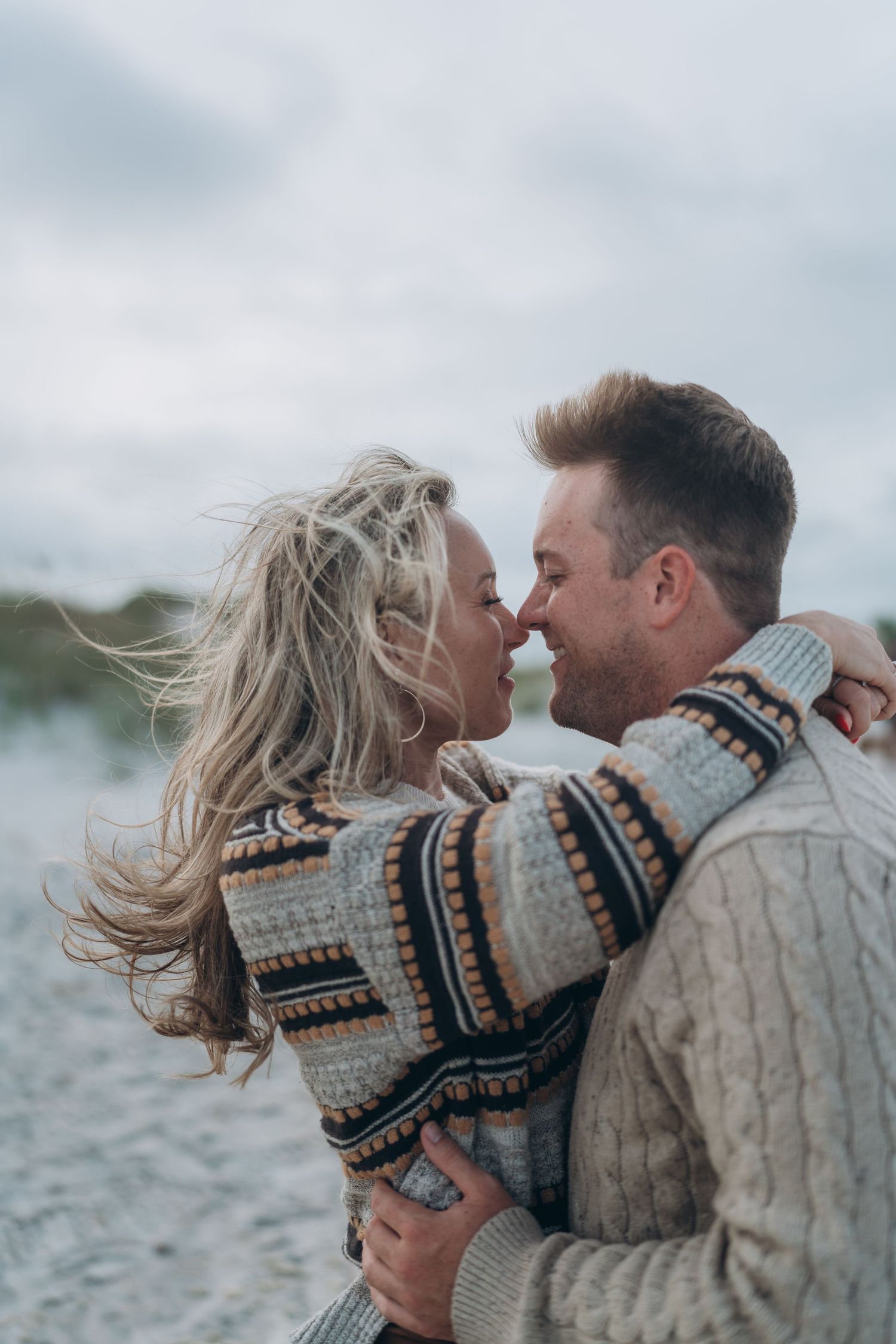 A couple in warm sweaters shares an intimate moment on a windy beach.