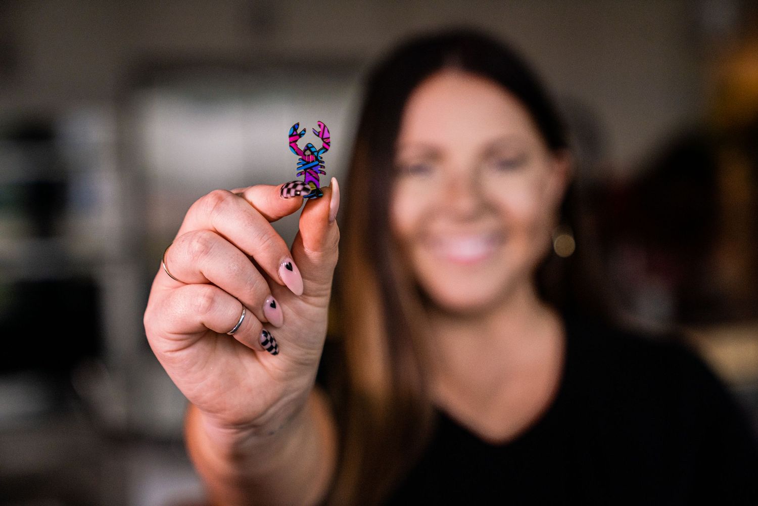 Smiling woman holding a tiny colorful 3D printed crab figurine between her fingers, with a blurred background.