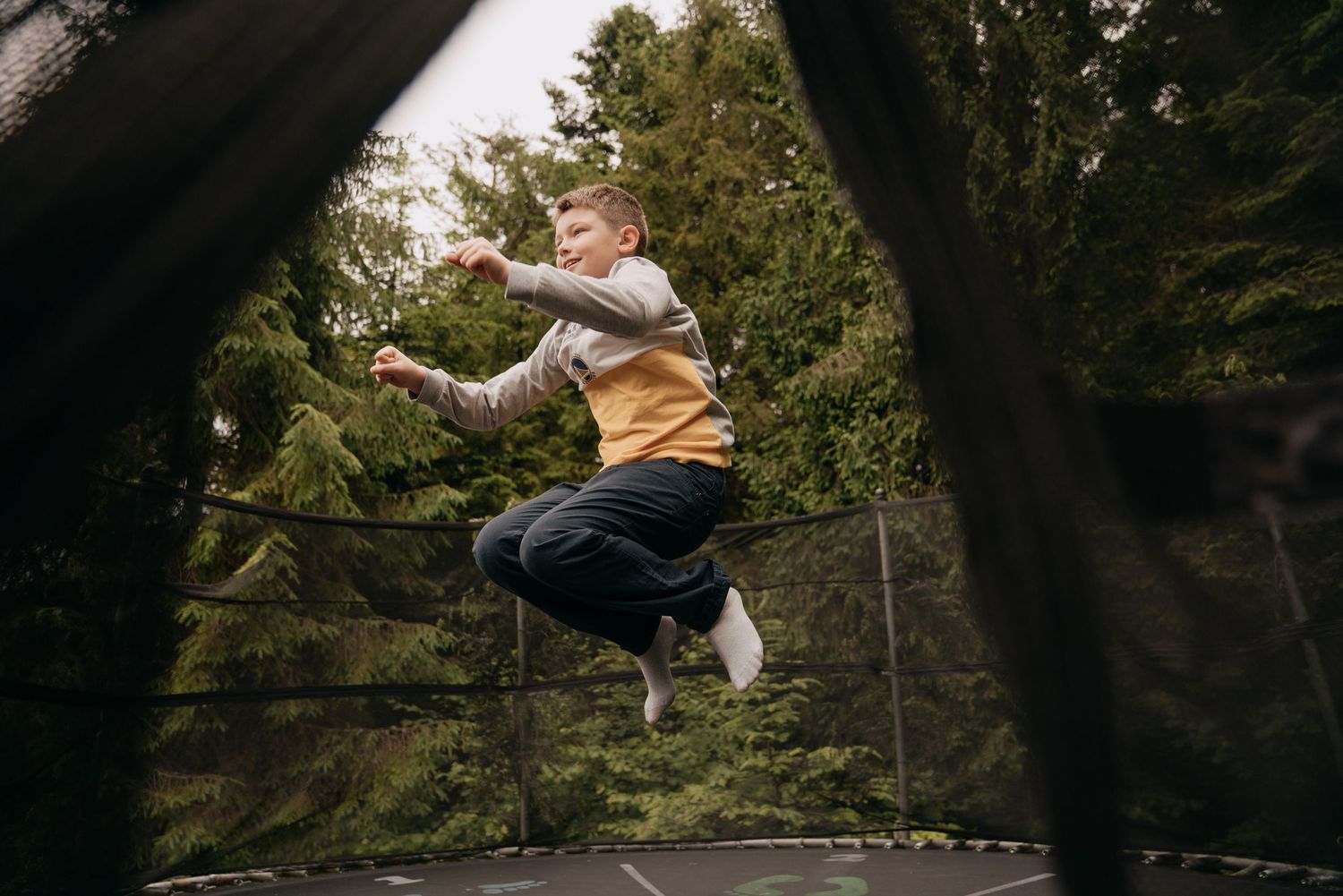 A young boy jumping high on a backyard trampoline surrounded by green trees.