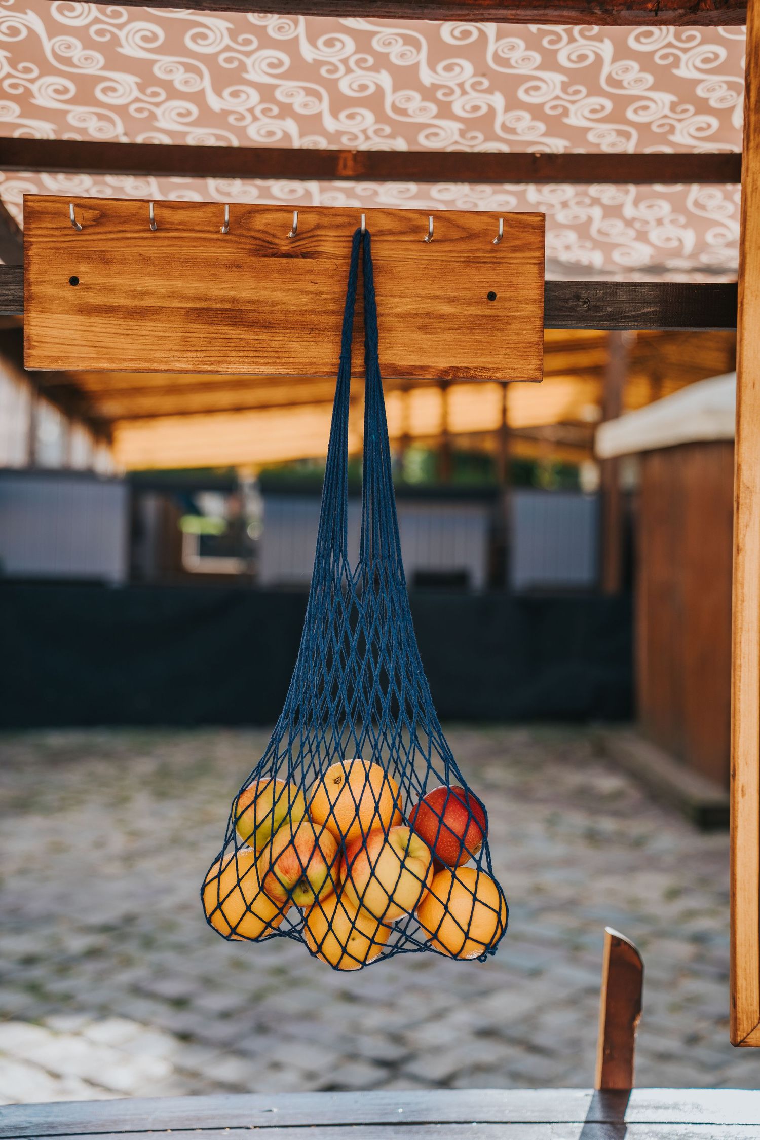 Blue mesh string bag filled with fresh apples hanging from a wooden hook at an outdoor market stall.