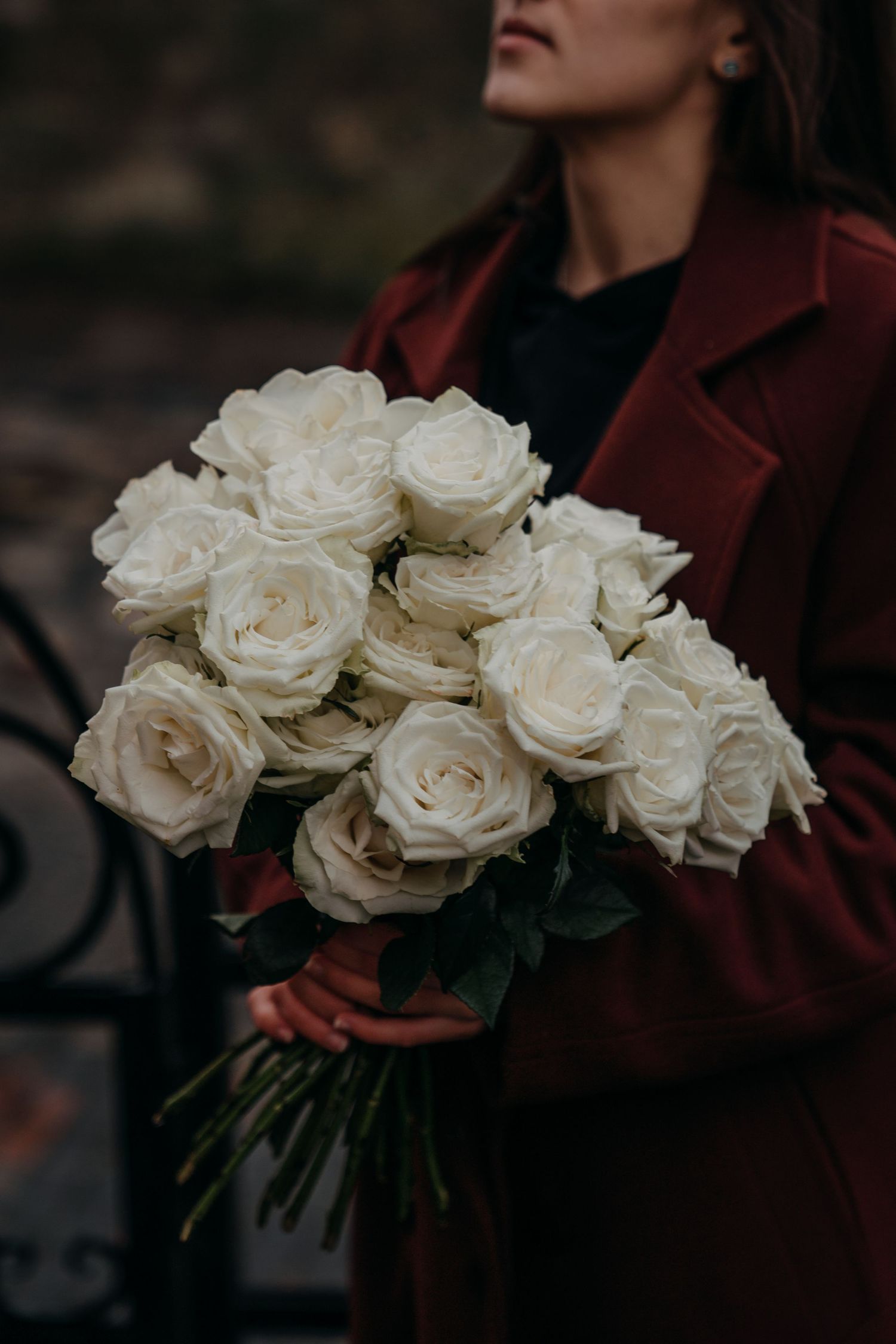 A woman in a red coat holds a large elegant bouquet of fresh white roses with green leaves outdoors.