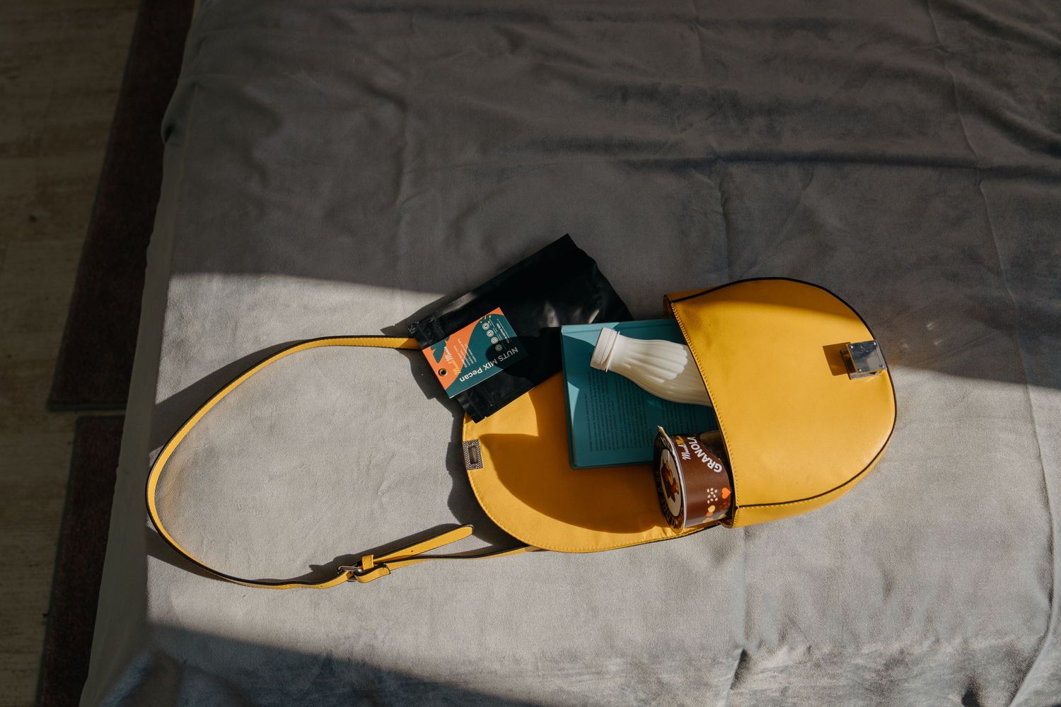 Yellow leather crossbody bag with teal wallet and granola snack bar viewed from above on gray fabric.