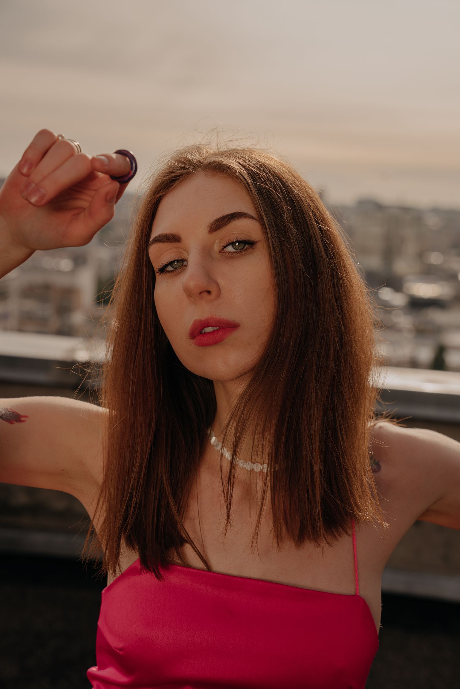 Young woman with brown hair in pink dress poses on rooftop at sunset, wearing necklace and holding small object.