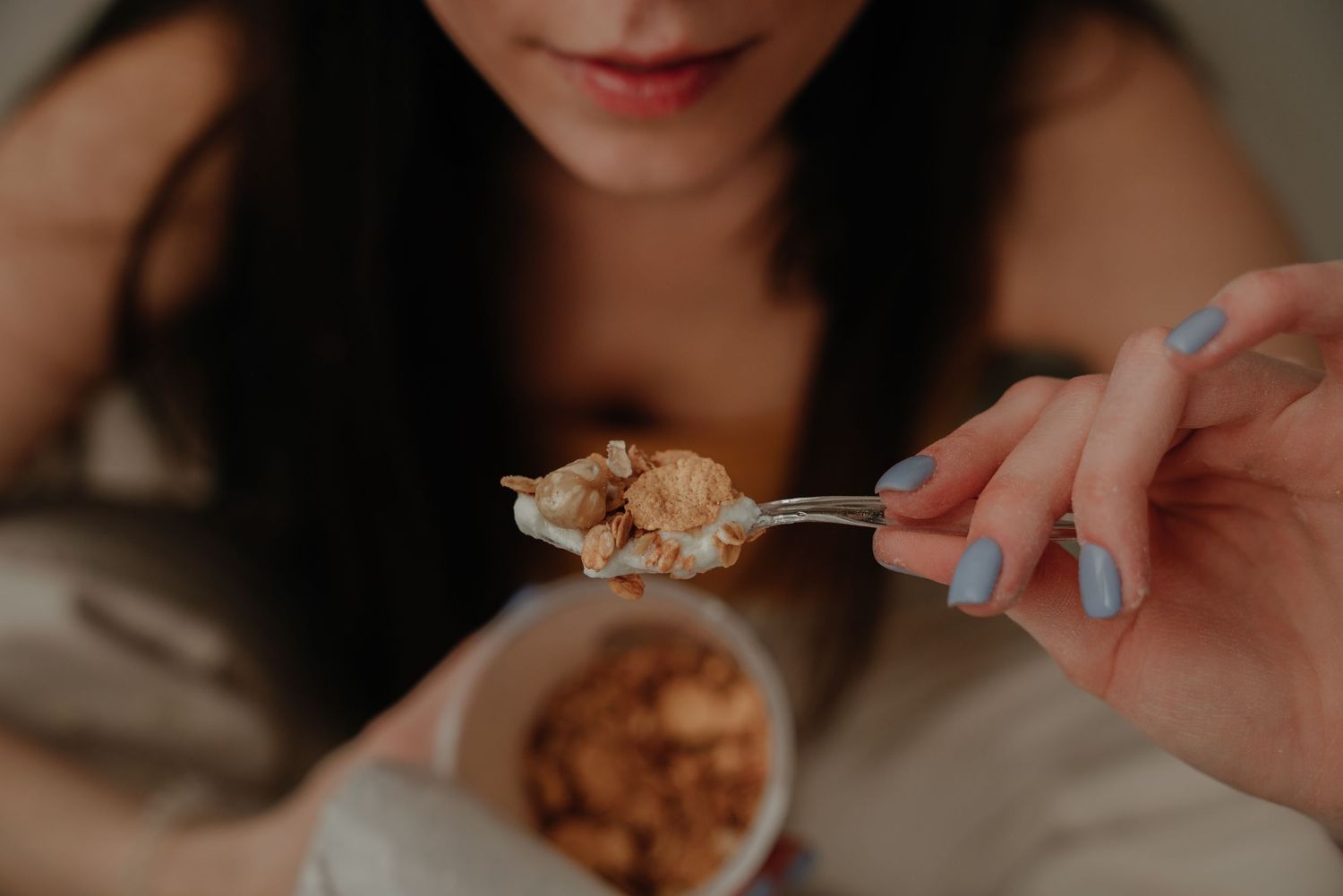 Woman with blue nails holding a spoon of granola over a bowl, enjoying a healthy snack.