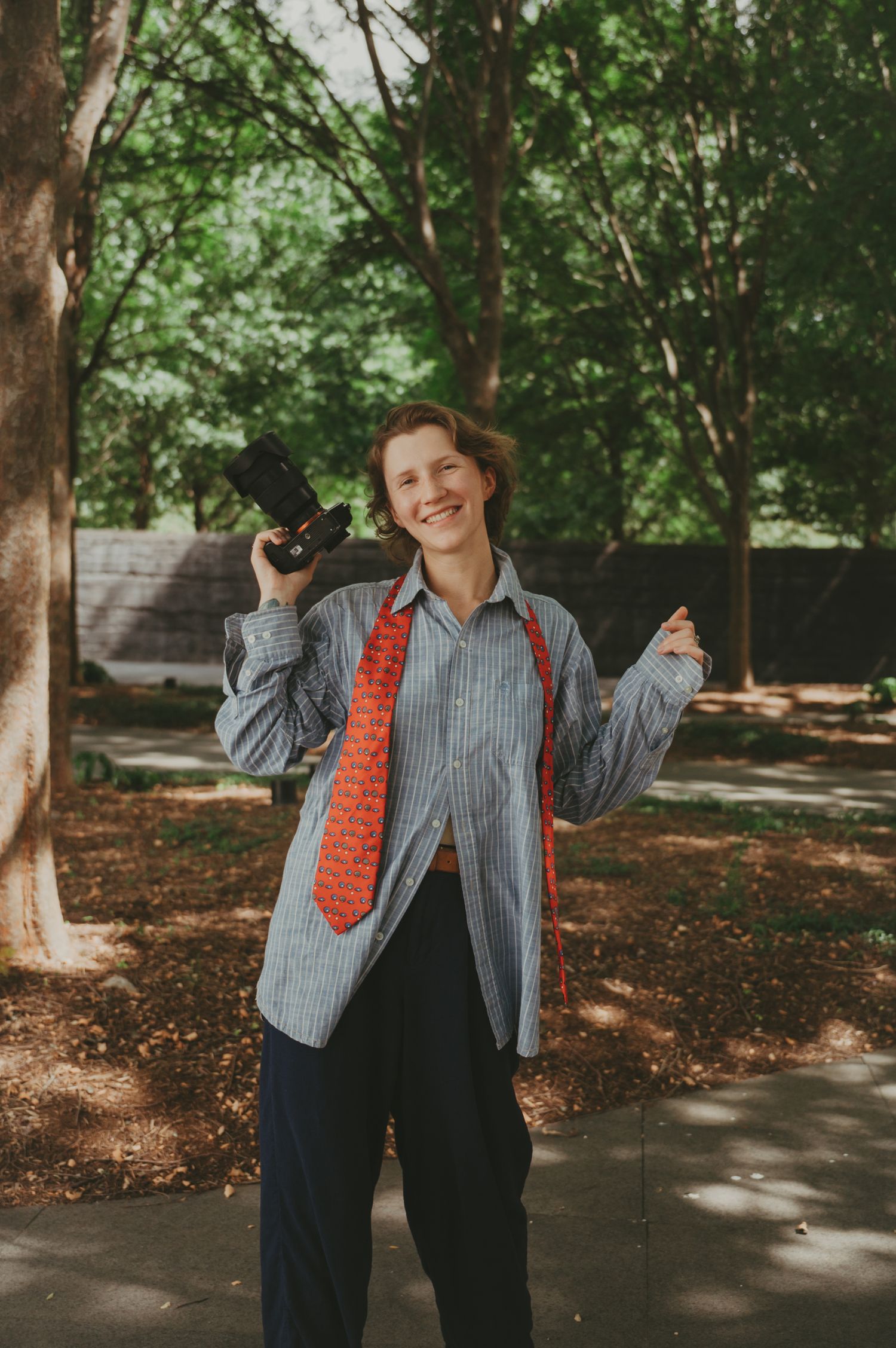 Person in striped shirt and red tie poses playfully in a wooded outdoor setting.