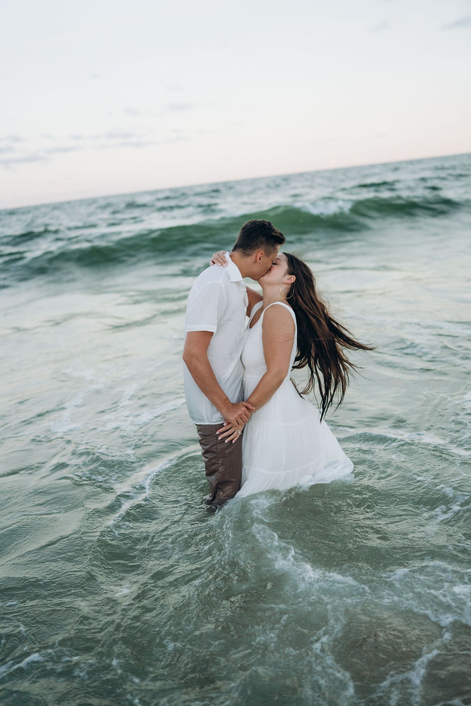 Couple kissing in ocean waves at sunset, woman in white dress and man in white shirt standing in shallow water.