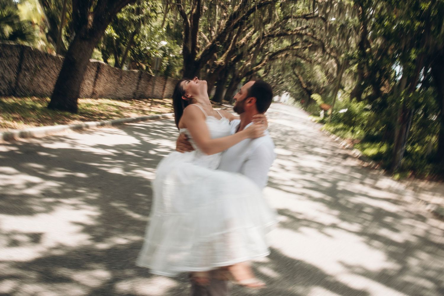 A groom joyfully lifts his bride in a white dress on a tree-lined path, spinning her in a romantic outdoor celebration.
