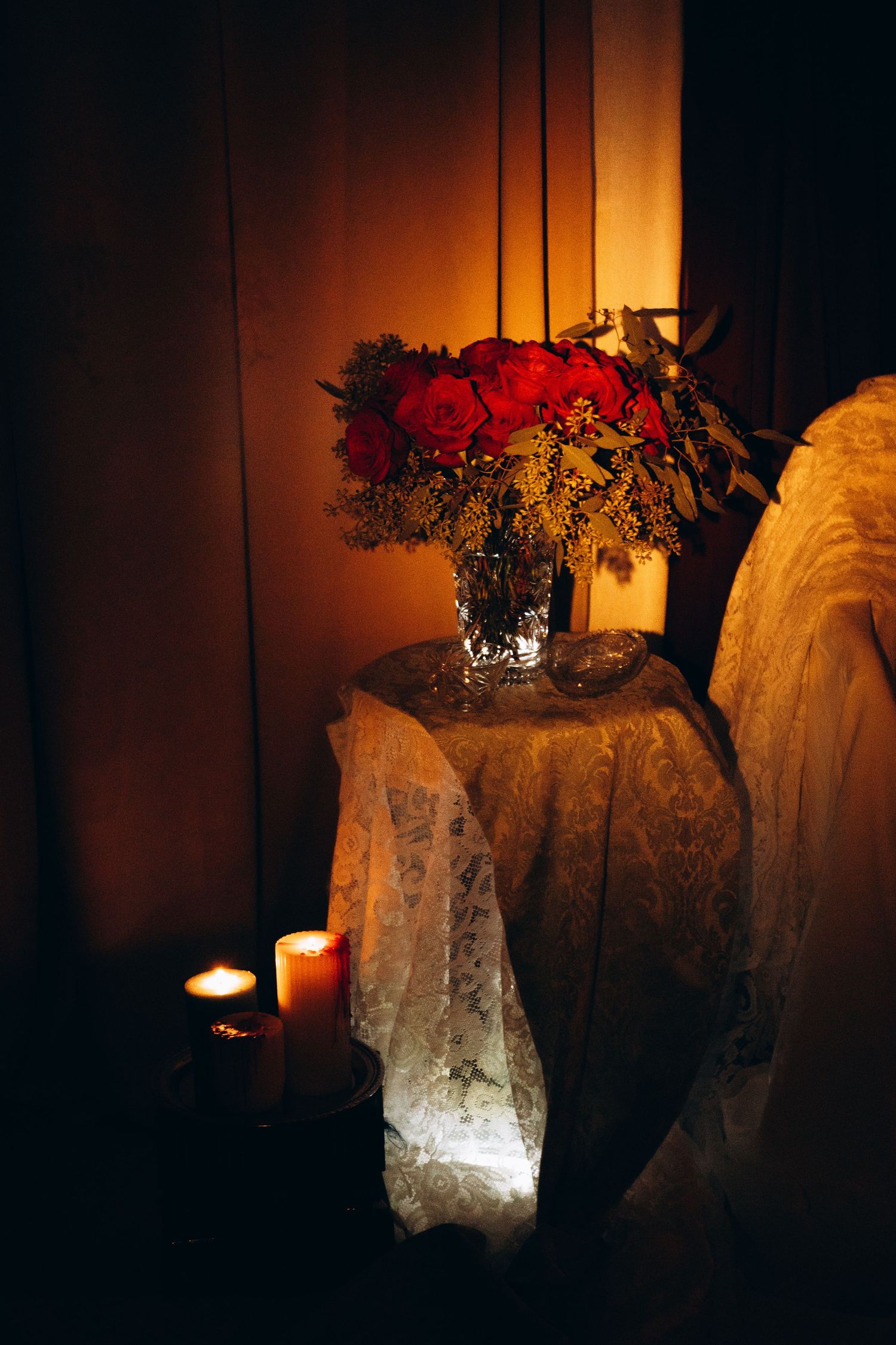 Crystal vase with red roses and candles on lace-covered table in warm candlelit romantic setting.